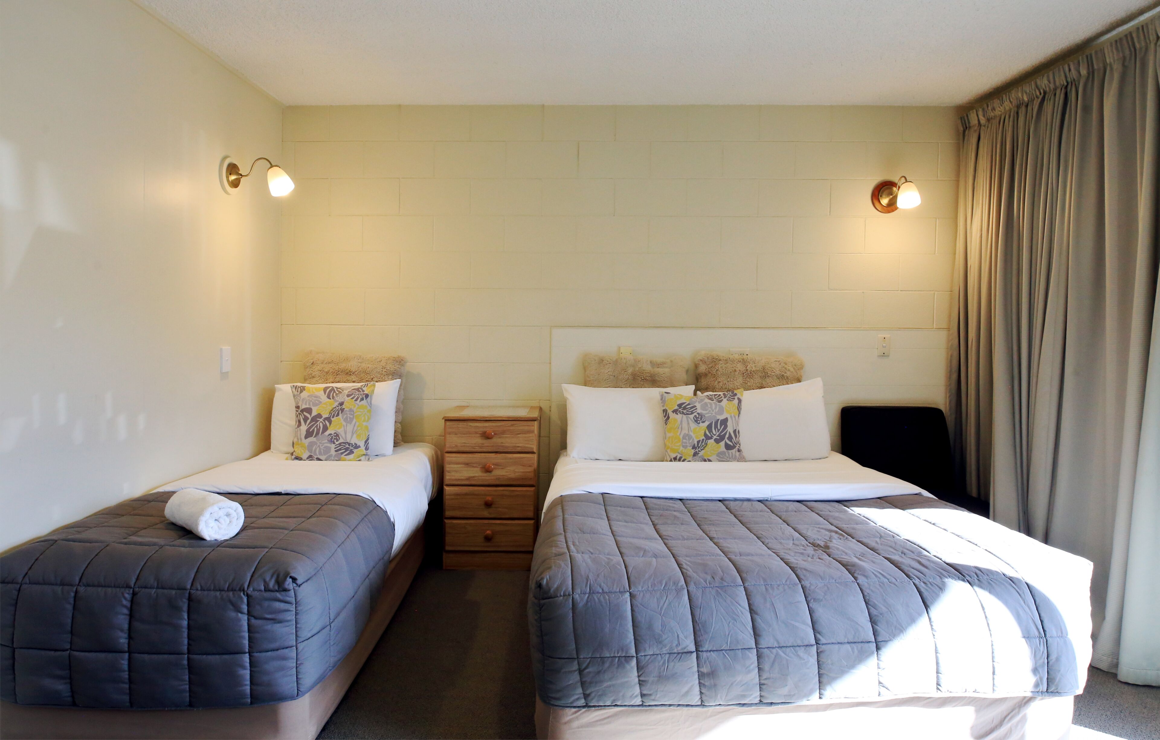A hotel room with two single beds, a wooden dresser, and a light-colored tiled wall.