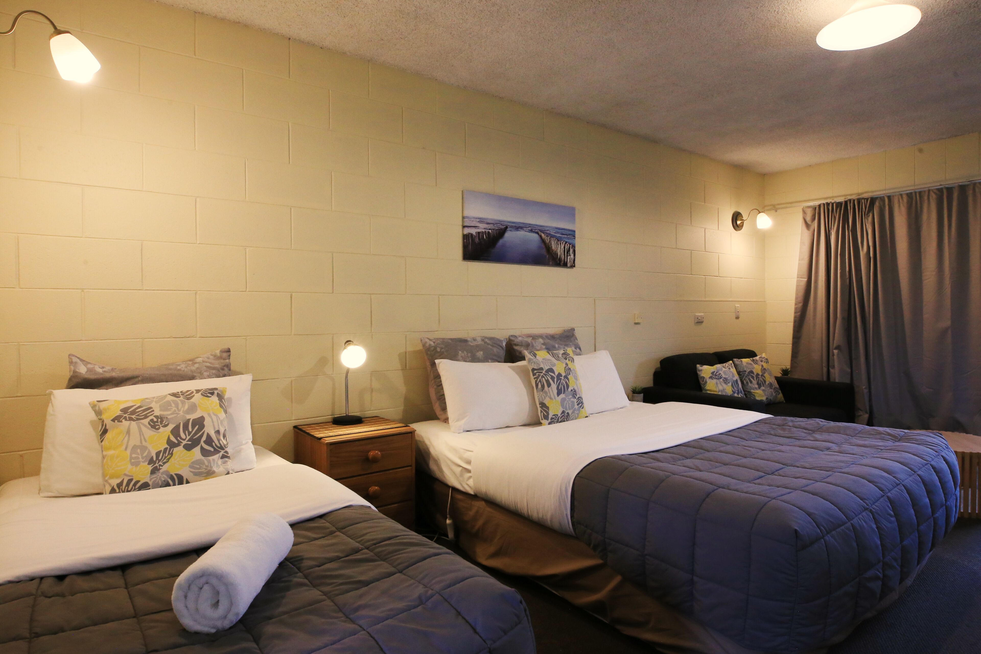 A hotel room with two beds, a nightstand, a lamp, a painting on the wall, and a curtain.
