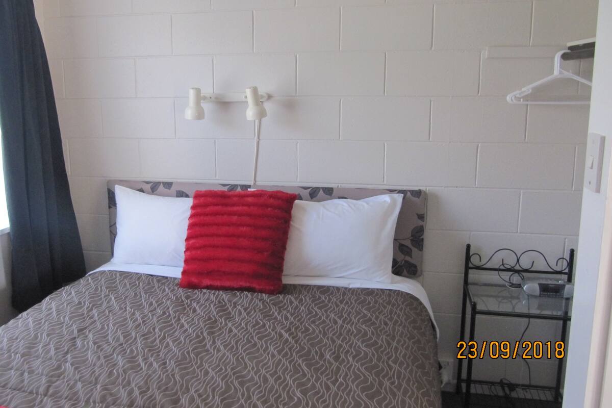 Quarto de hotel com uma cama, janela com cortinas, luminária fixada na parede e uma pequena mesa com um copo.