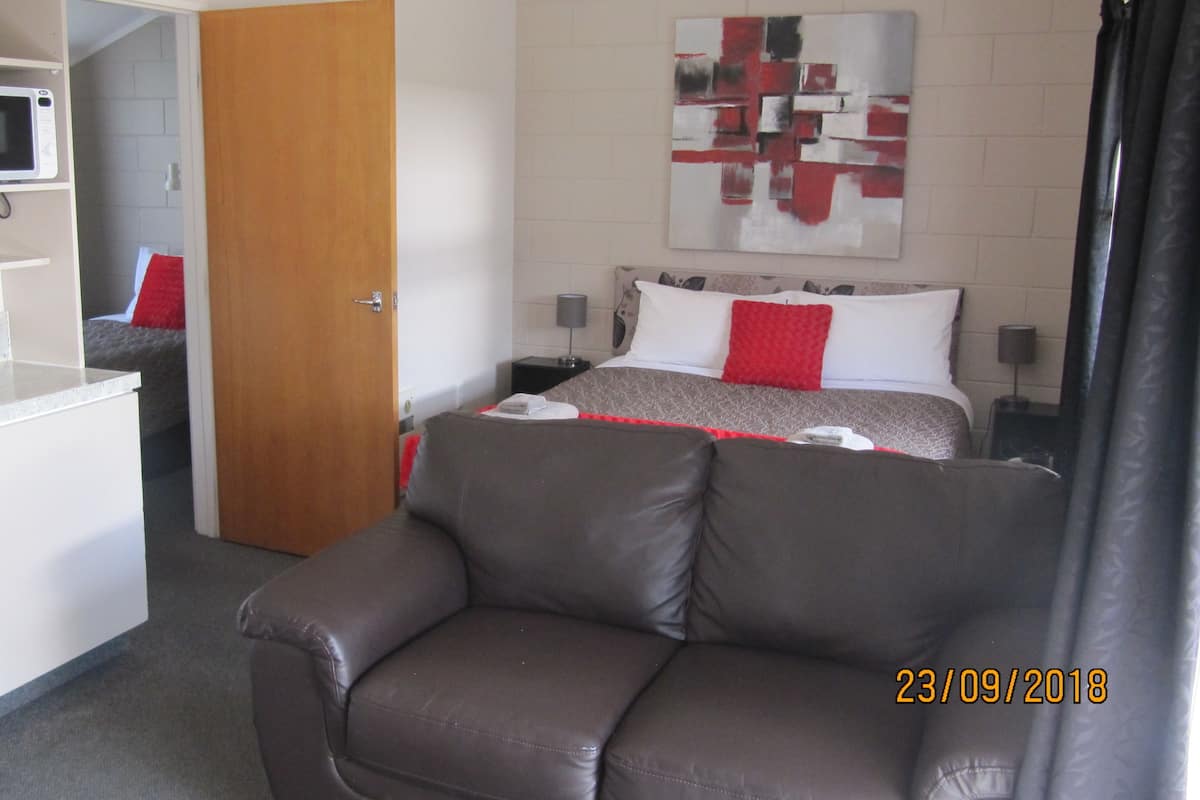 Quarto de hotel compacto com cozinha compacta, cama com roupa de cama vermelha e branca, sofá cinza e uma pintura abstrata moderna na parede.