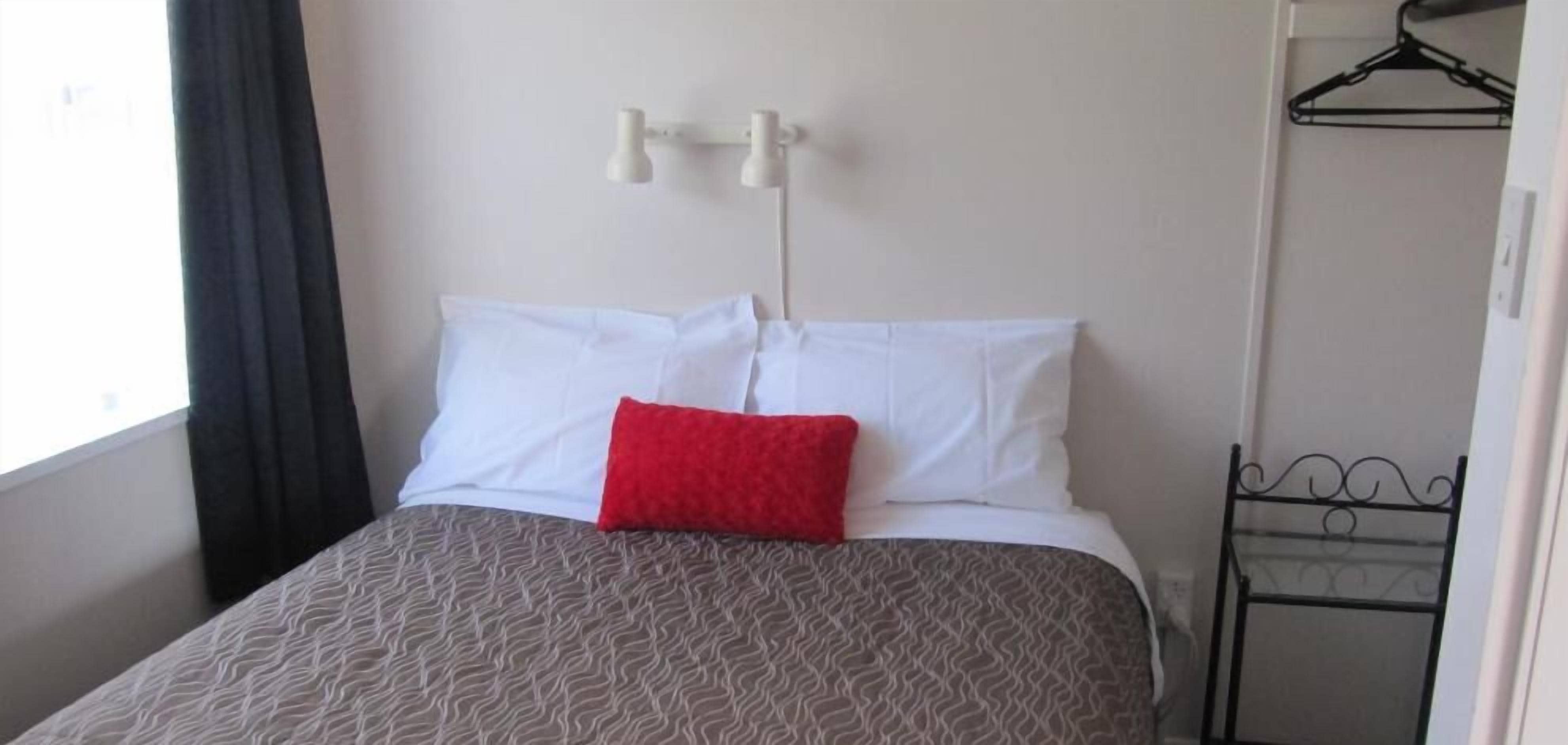 A neatly made bed with a patterned bedspread and a single red pillow, a white wall with a simple light fixture, and a black metal rack against the wall.
