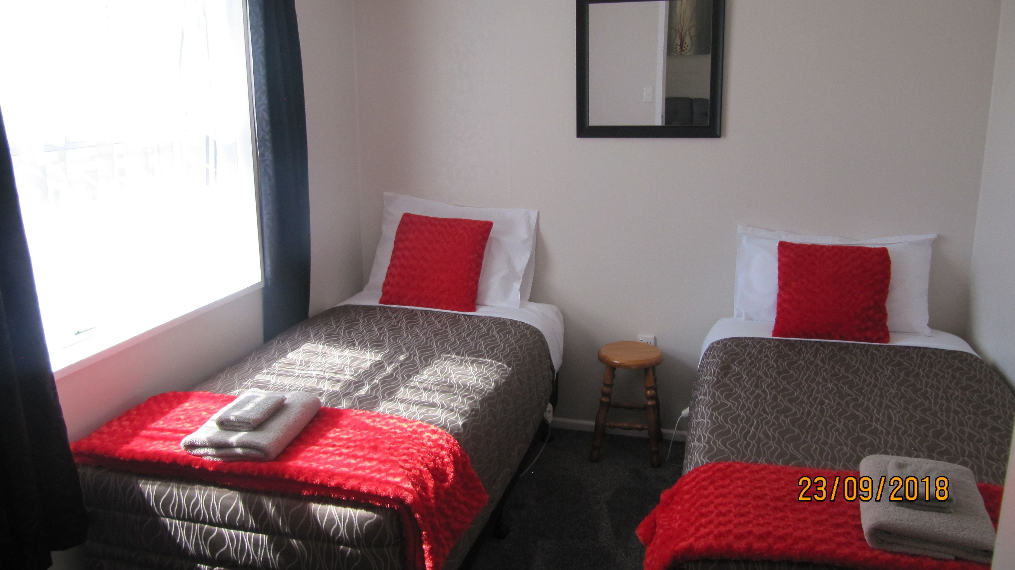 Habitación de hotel con dos camas, cada una con una colcha estampada y almohadas rojas. Hay un taburete pequeño y un espejo en la pared.