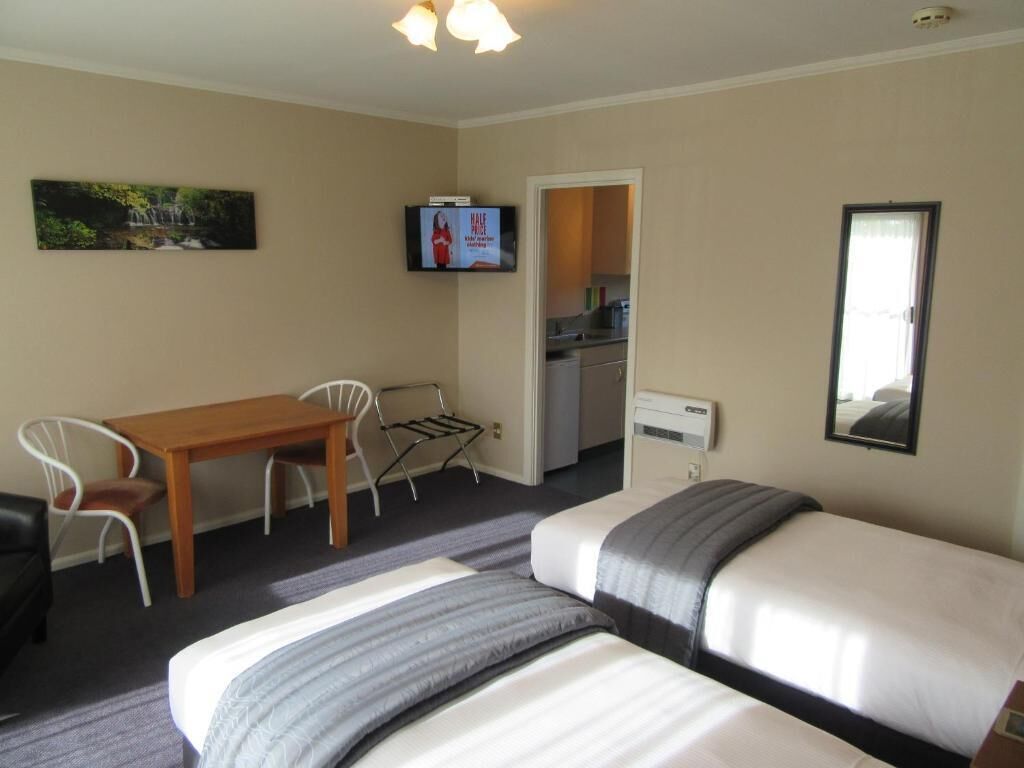 A hotel room with two beds, a wooden table, a TV, and a kitchen area visible through an open door.