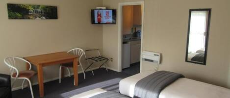 A hotel room with two beds, a wooden table, a TV, and a kitchen area visible through an open door.