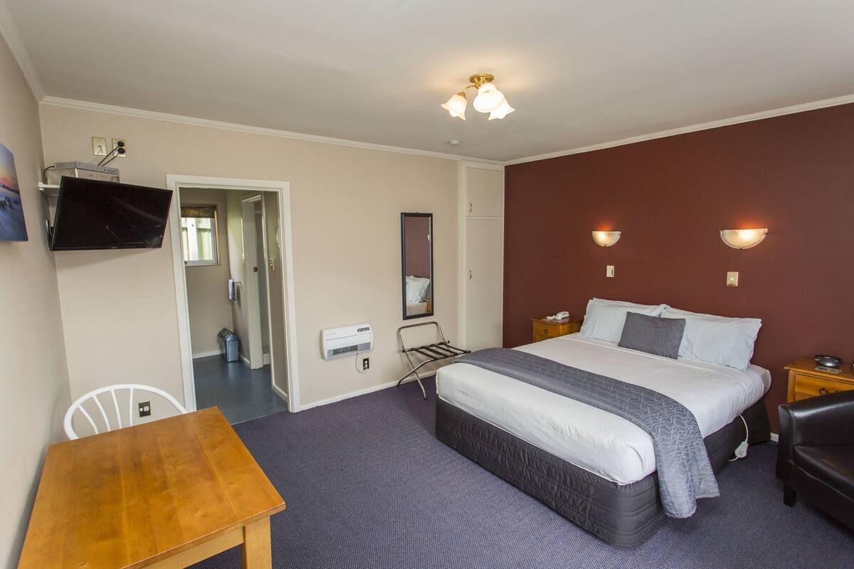 A hotel room with a bed, bedside table, chair, TV, and a mirror.