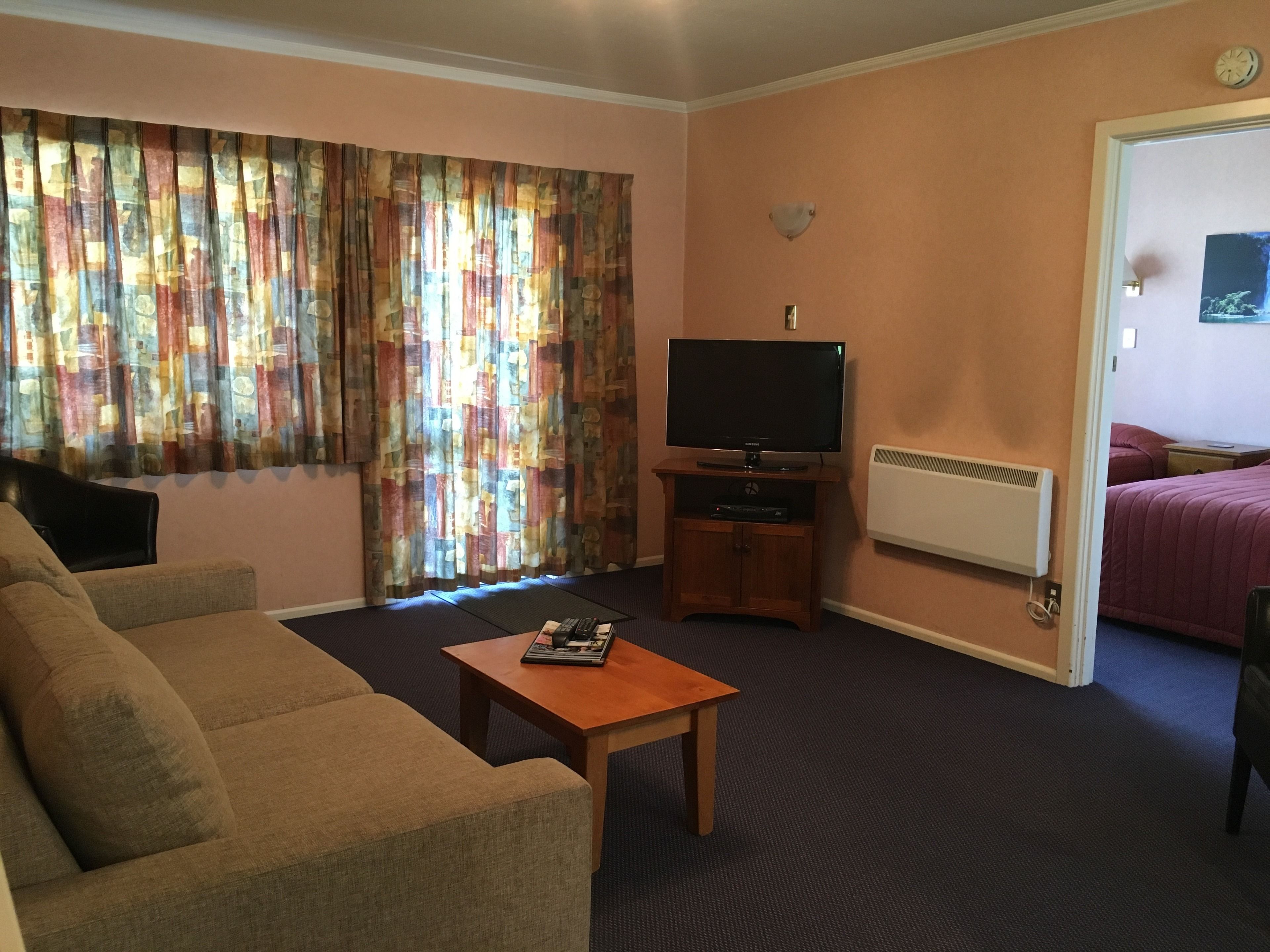 Una sala de estar con sofá, una mesa de centro, un televisor de pantalla plana sobre un soporte de madera, una ventana con cortinas, y una cama en la habitación contigua.