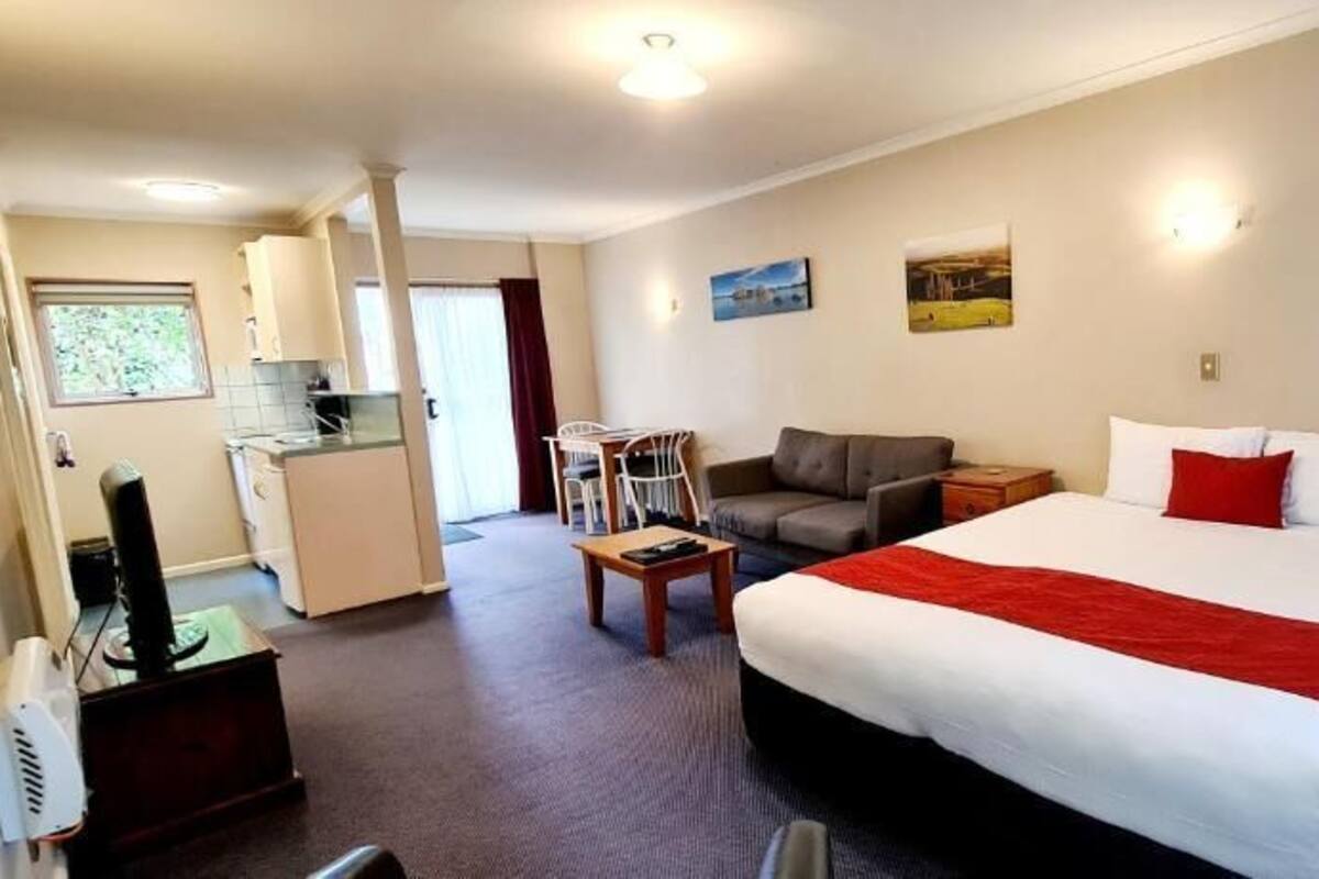 A hotel room with a large bed, a desk, a chair, a sofa, a small kitchen area, and a dining table.