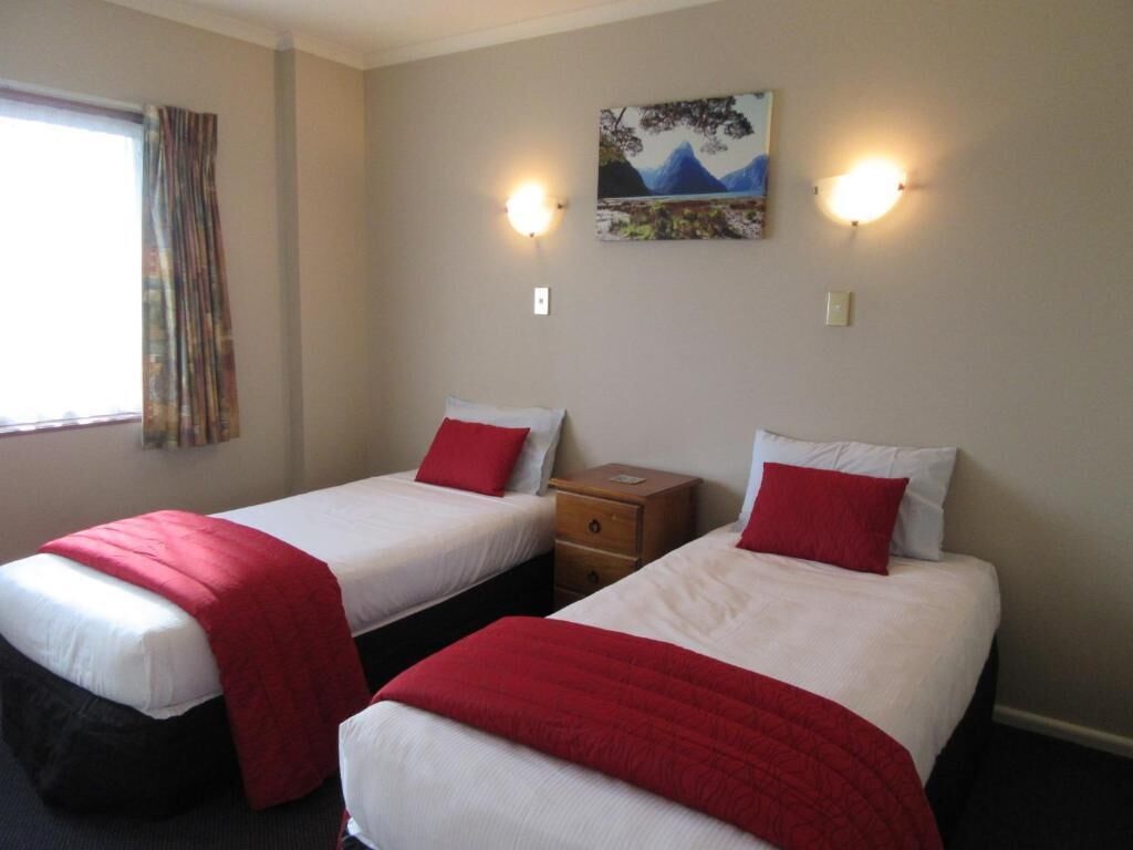 A hotel room with two beds, each with a red blanket and white pillows. There is a painting on the wall and two wall-mounted lights.