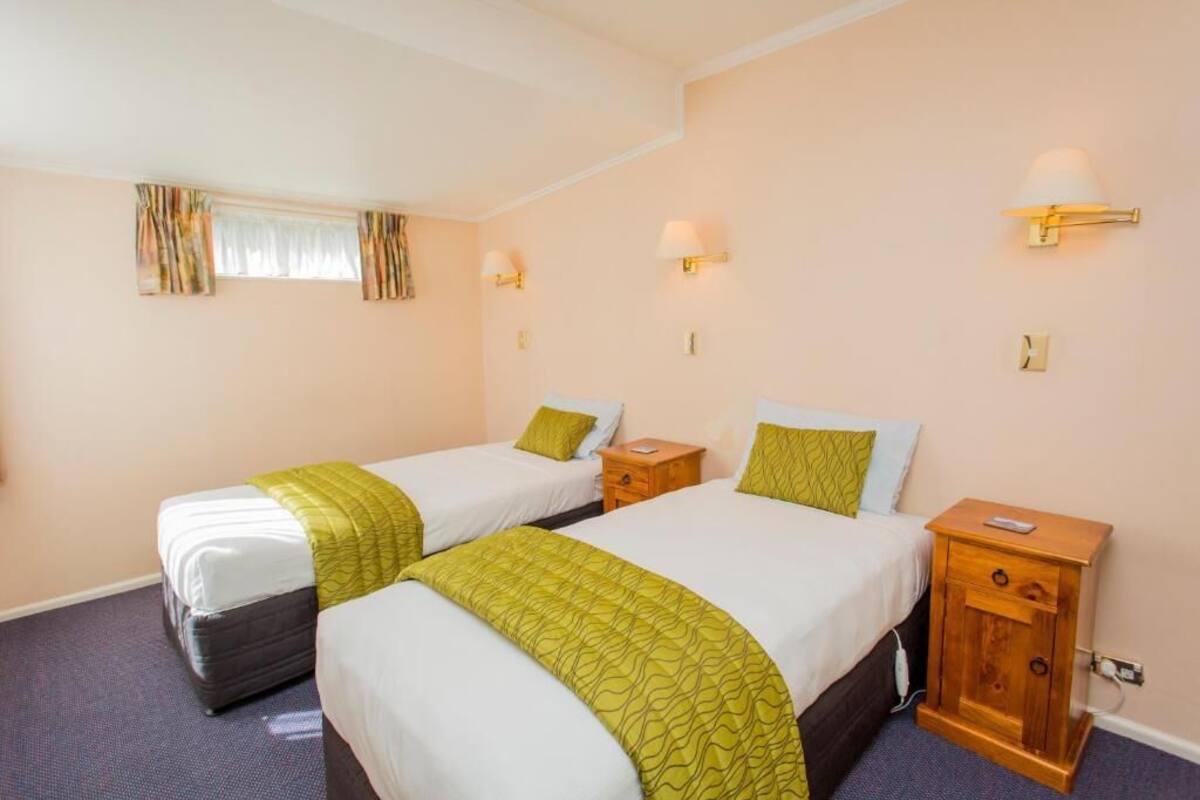 A hotel room with two single beds, each with a green throw blanket, a wooden nightstand with a lamp, and a window with curtains.