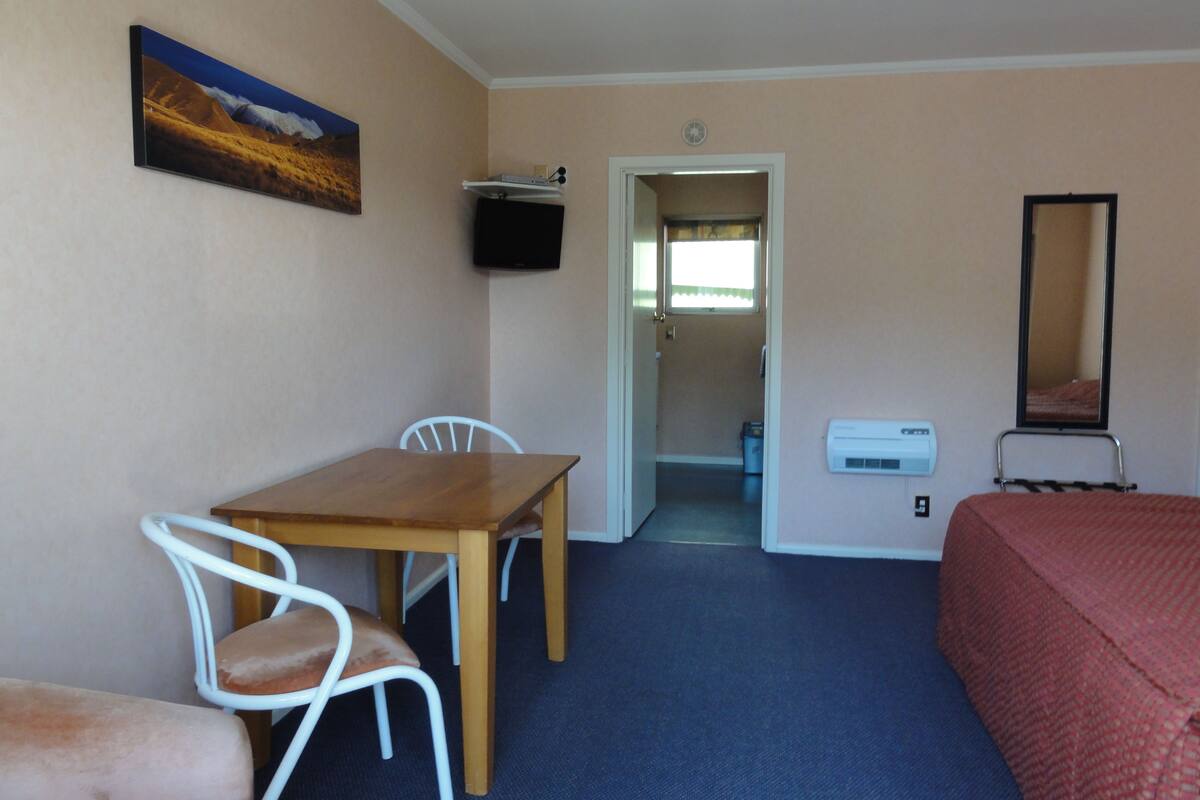 A room with a wooden table, two white chairs, a bed, a television, and a mirror.