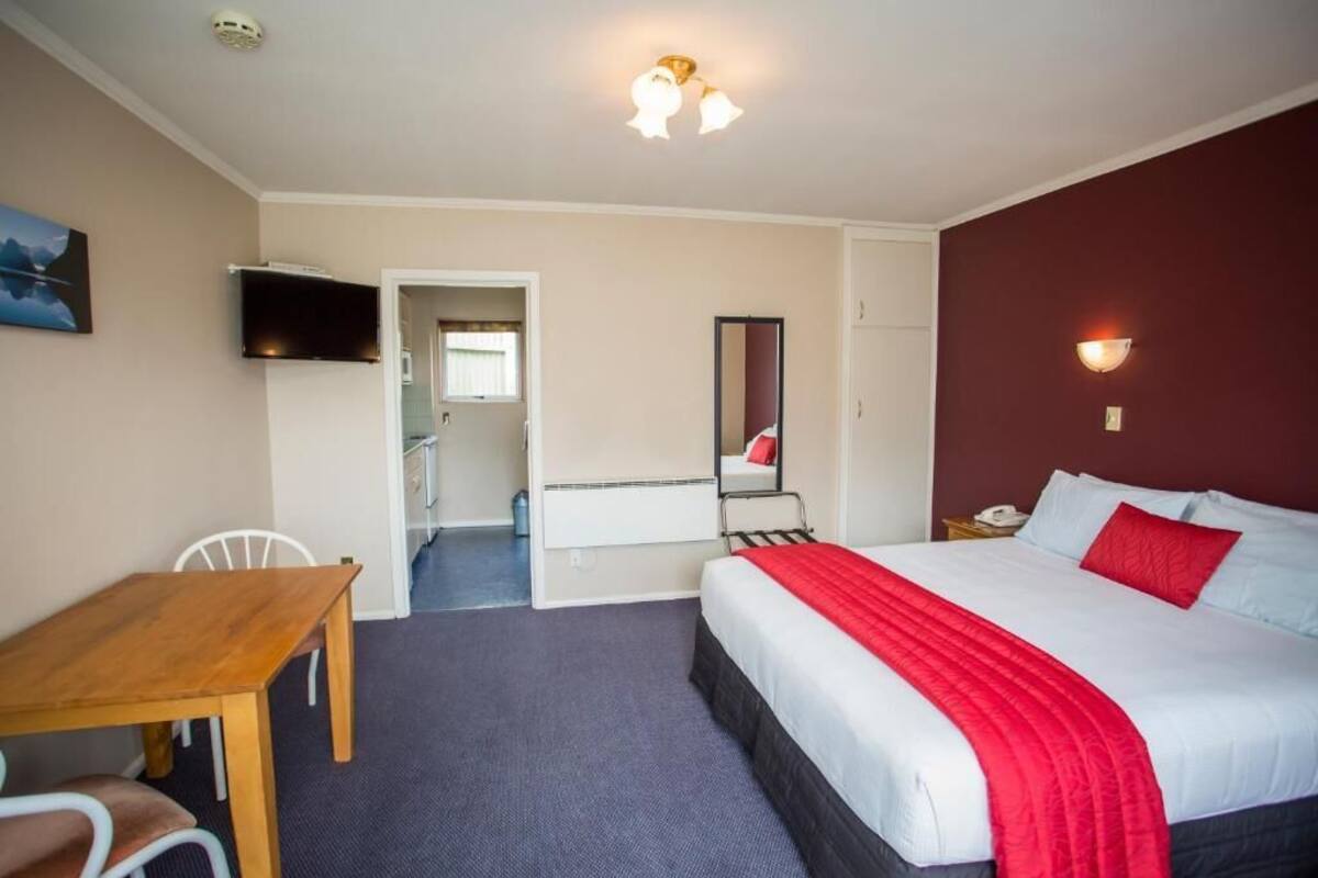 A hotel room with a bed, a desk, a chair, a TV, a mirror, and a door leading to another room.