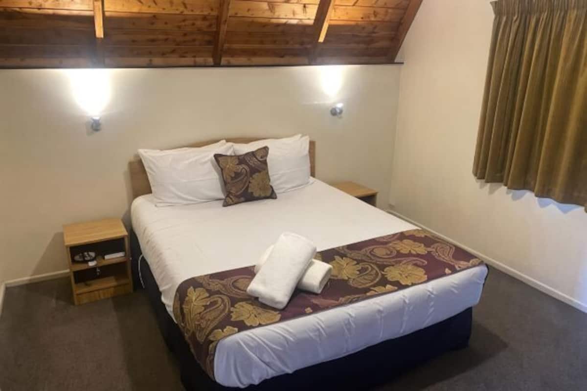 Un dormitorio con cama, dos mesitas de noche, un velador, una ventana con cortinas y techo de madera.