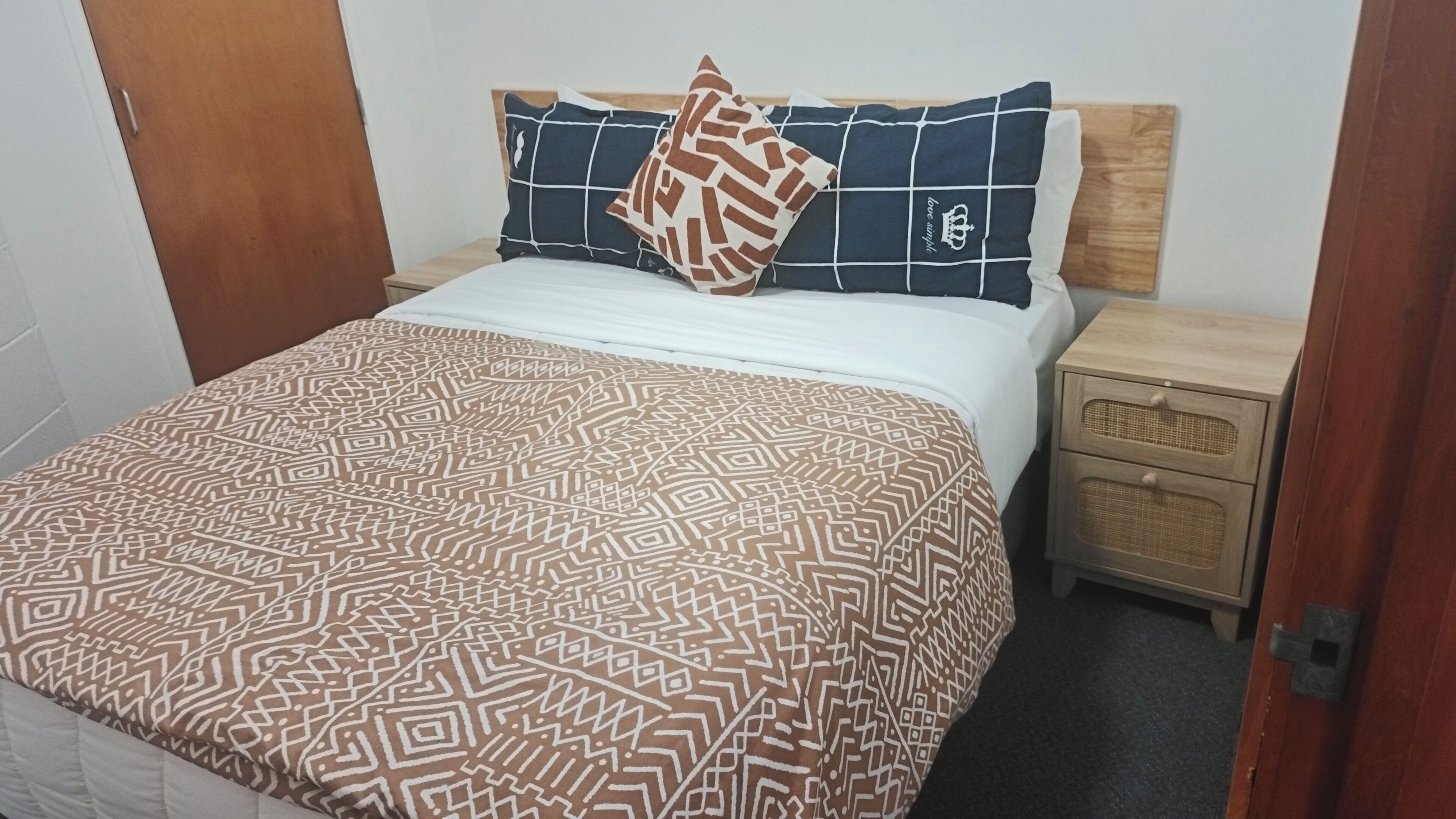 A bed with a patterned bedspread, a pillow with a geometric design, and a matching pillow with a grid pattern. There is a wooden nightstand with a wicker basket drawer.