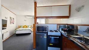 Une chambre d’hôtel compacte comprenant un coin cuisine, un lit avec une literie à motifs floraux, une télévision et une petite salle de bain.