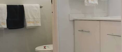 Un baño con inodoro, lavamanos y espejo. Hay toallas colgadas en la pared y una puerta que da a otra habitación.