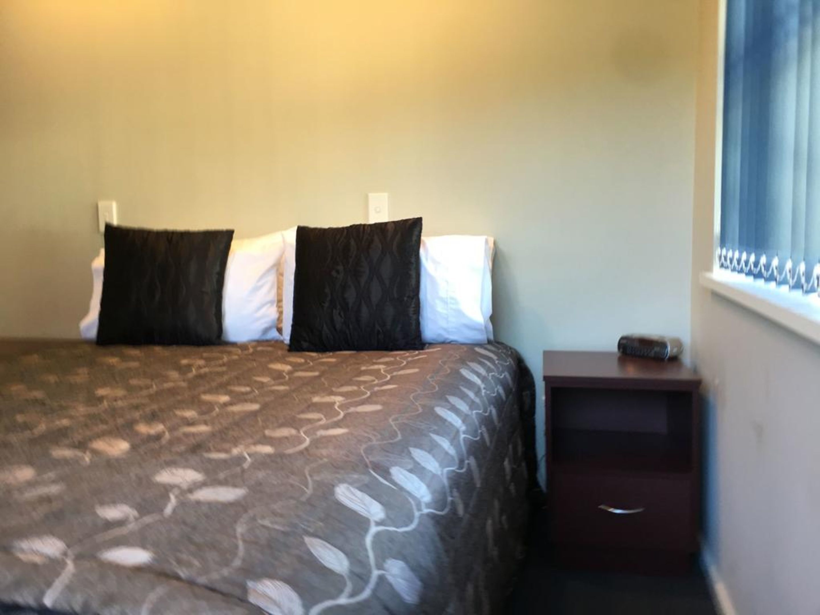 Un dormitorio con cama, dos almohadas, una mesita de noche y una ventana con cortinas.