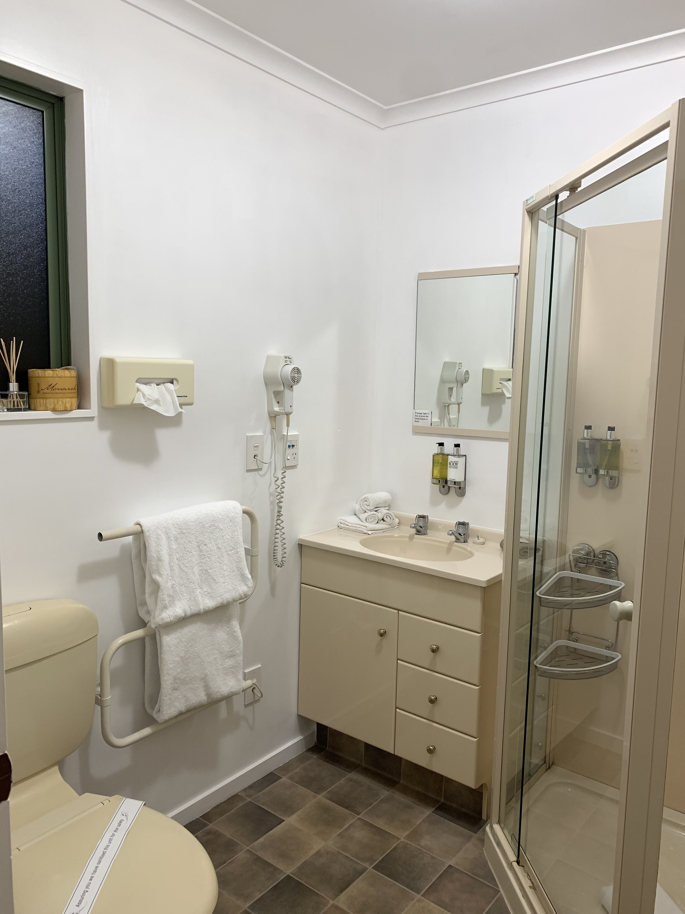 Un baño con inodoro, lavamanos y ducha. Cuenta con un perchero, un espejo y un box de ducha de vidrio.