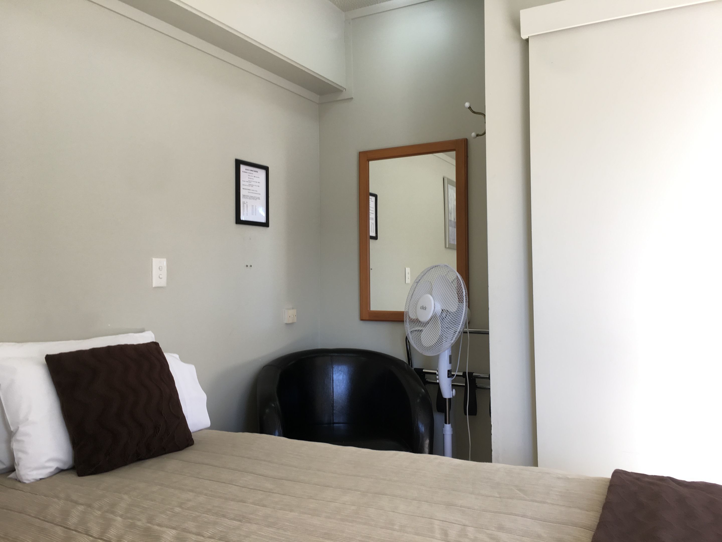 A hotel room with a bed, a chair, a standing fan, and a mirror on the wall.