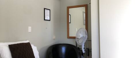 A hotel room with a bed, a chair, a standing fan, and a mirror on the wall.