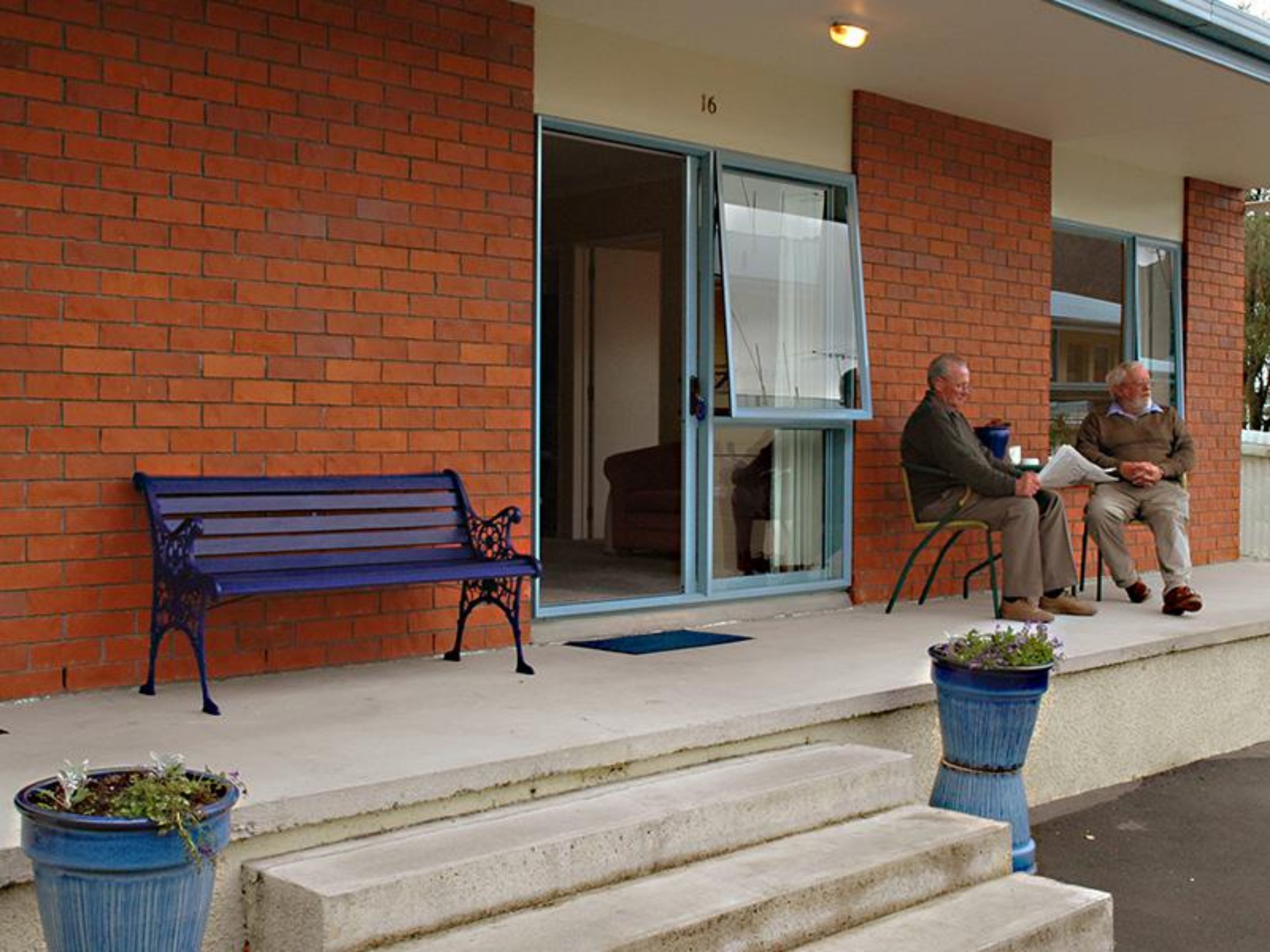 A brick building with a blue bench outside, a glass door, and two potted plants.