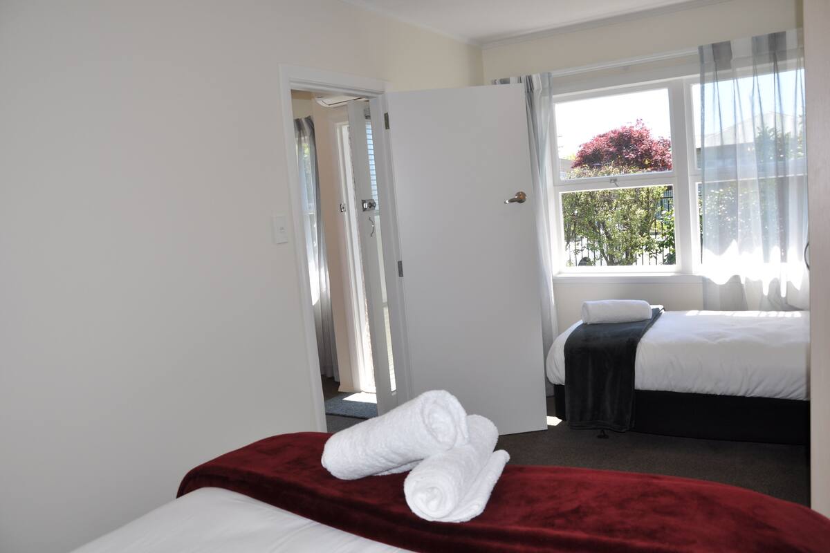 Una habitación de hotel con cama, una colcha roja y una ventana con cortinas.