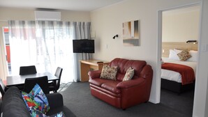 A hotel room with a leather sofa, a desk with a flat-screen TV, a bed with a red blanket, and a painting on the wall. - Balmoral Motel (Nelson)