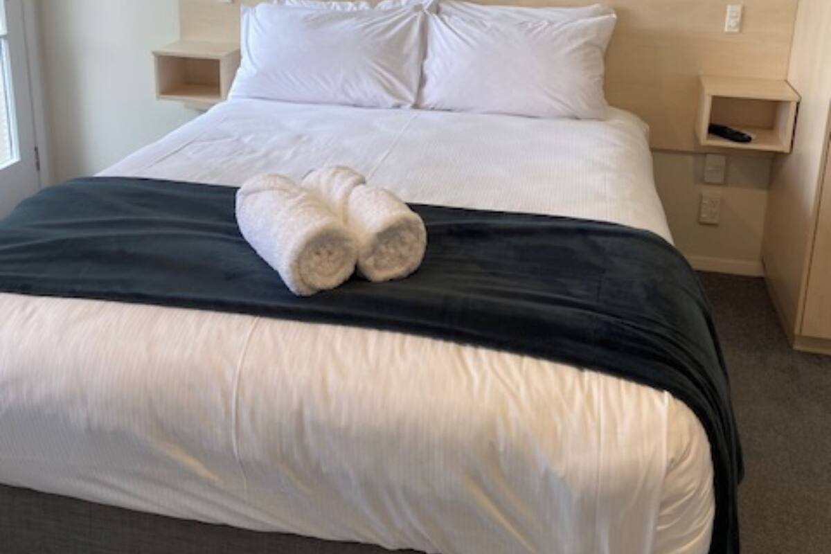 Una cama bien hecha con sábanas blancas y un plaid negro, junto con dos toallas enrolladas sobre la cama.