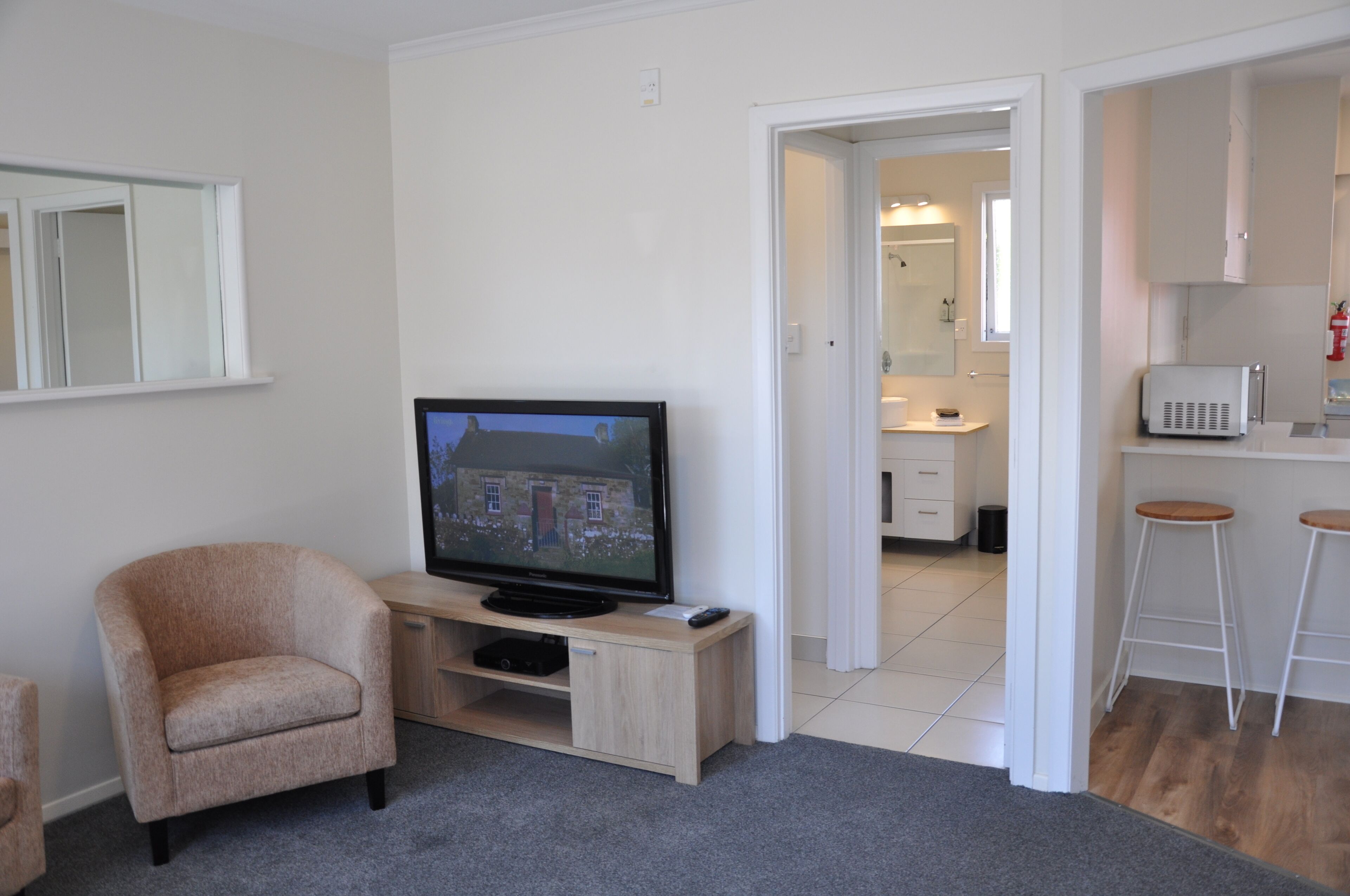 Una sala de estar con televisor sobre un mueble de madera, un sillón color beige y una zona de cocina con taburetes y armarios.