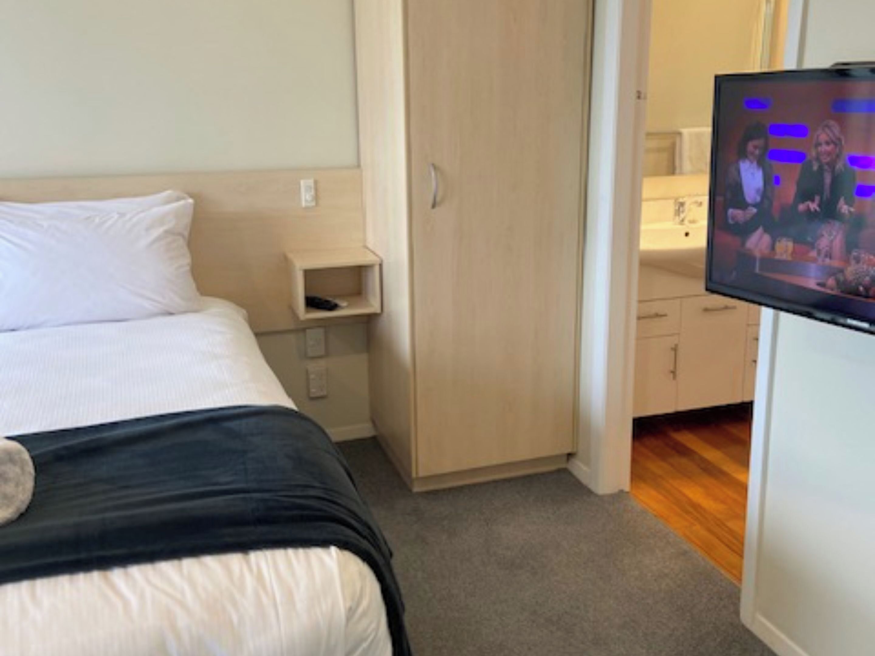Ein Hotelzimmer mit Bett, Wandfernseher, Holzschrank und einem kleinen Nachttisch.