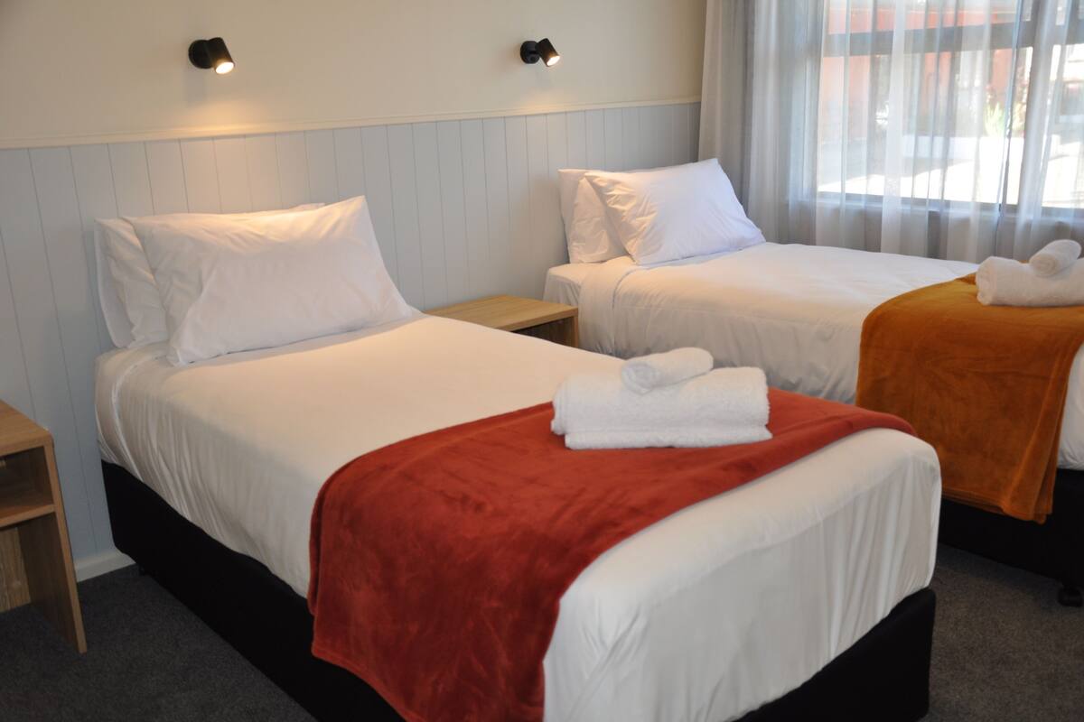 Habitación de hotel con dos camas, cada una con sábanas blancas y una colcha roja en la cama de la izquierda. Hay dos mesitas de noche con lámparas, y una ventana con cortinas traslúcidas.