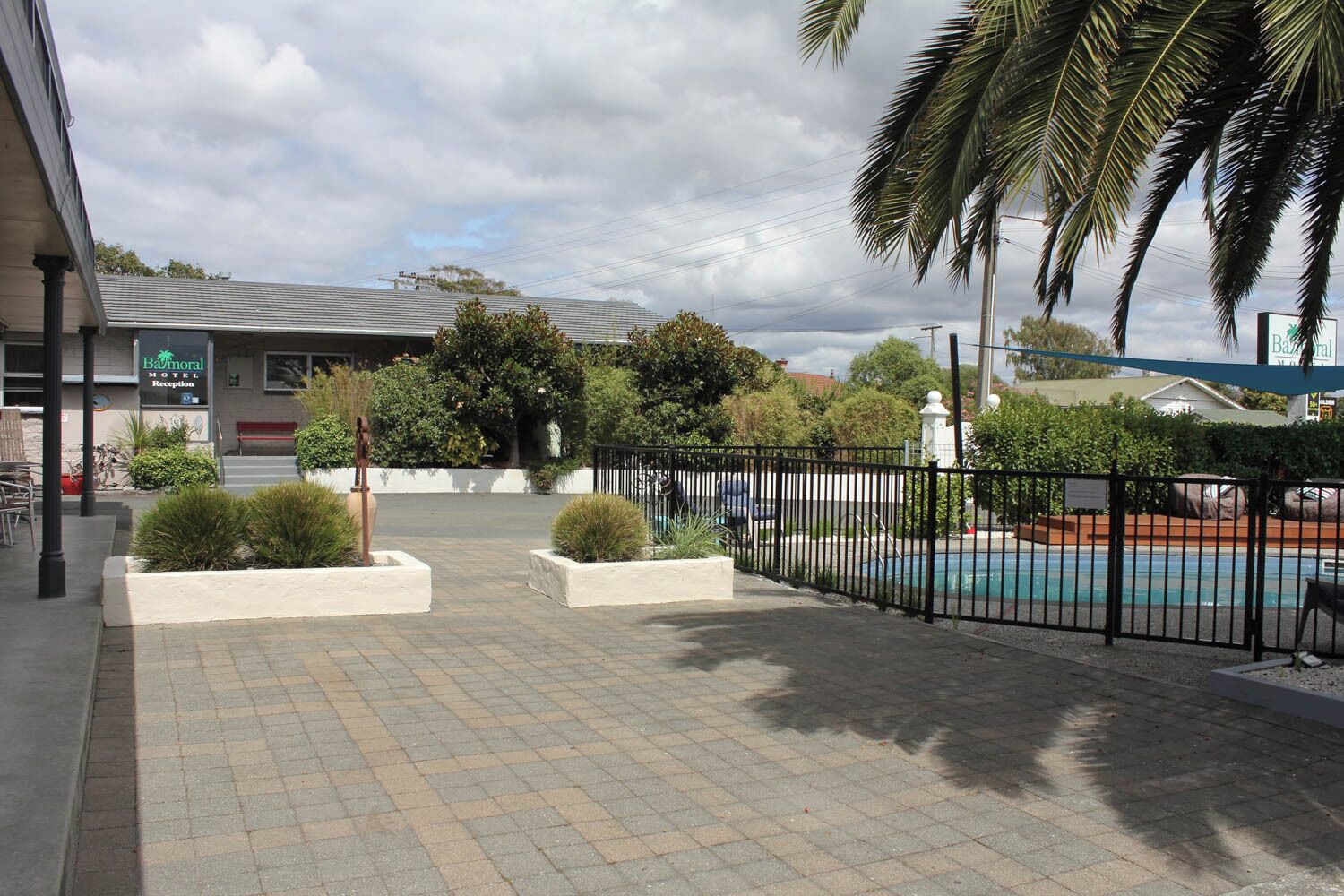 A motel with a reception desk, a fenced swimming pool, and landscaped areas with small bushes and palm trees.