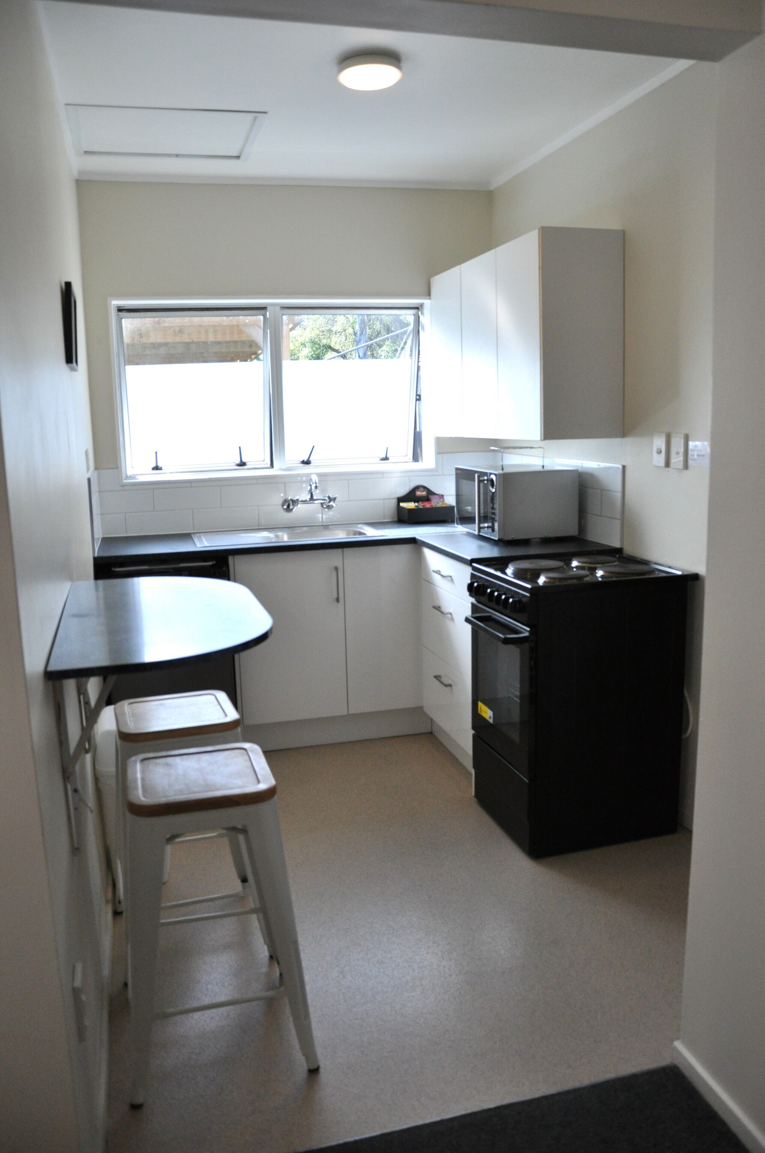 A compact kitchen with a sink, stove, microwave, and a small dining area with a stool.