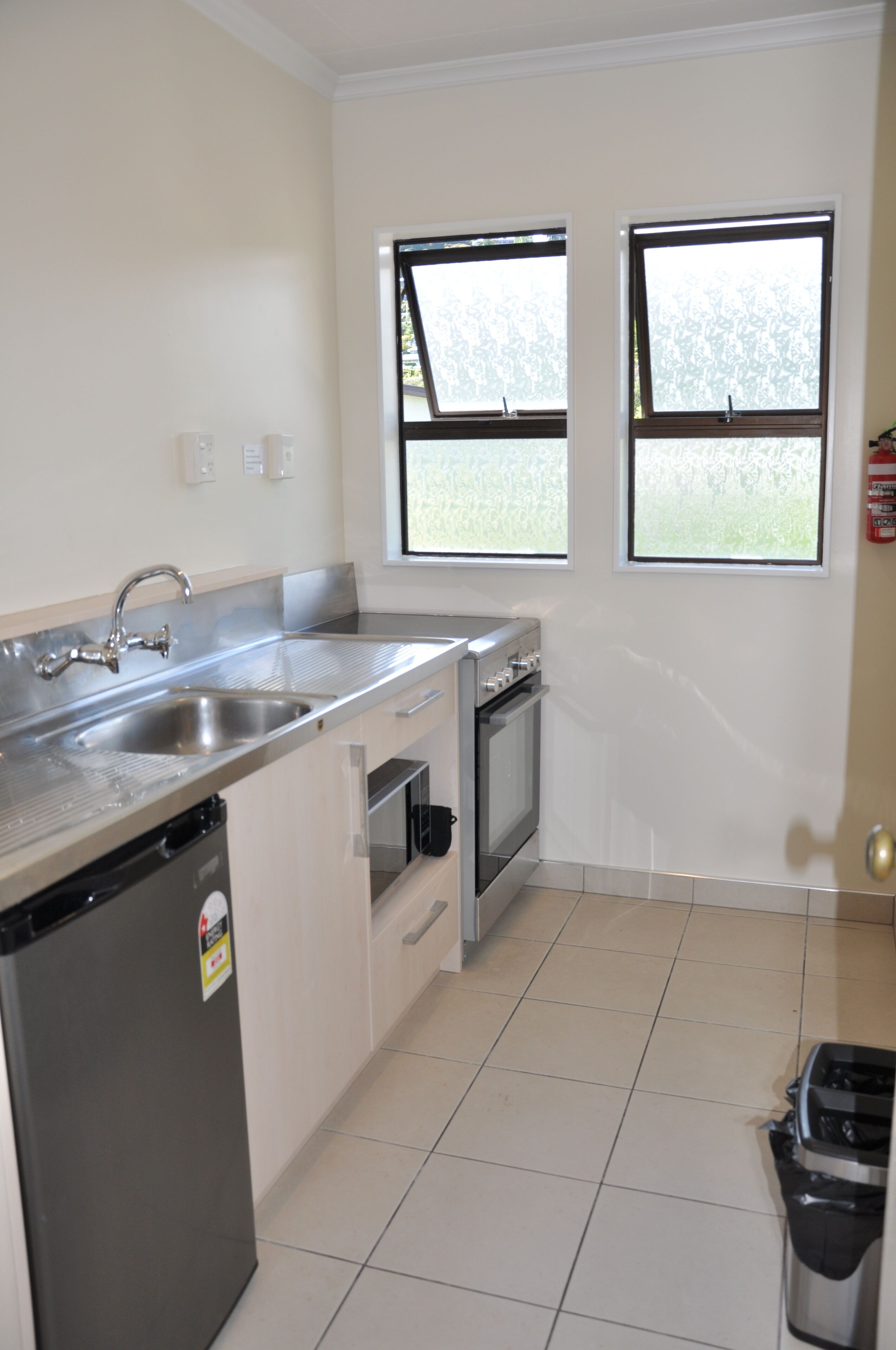 A compact kitchen with a stainless steel sink, a built-in oven, and a dishwasher. There are two windows providing natural light.