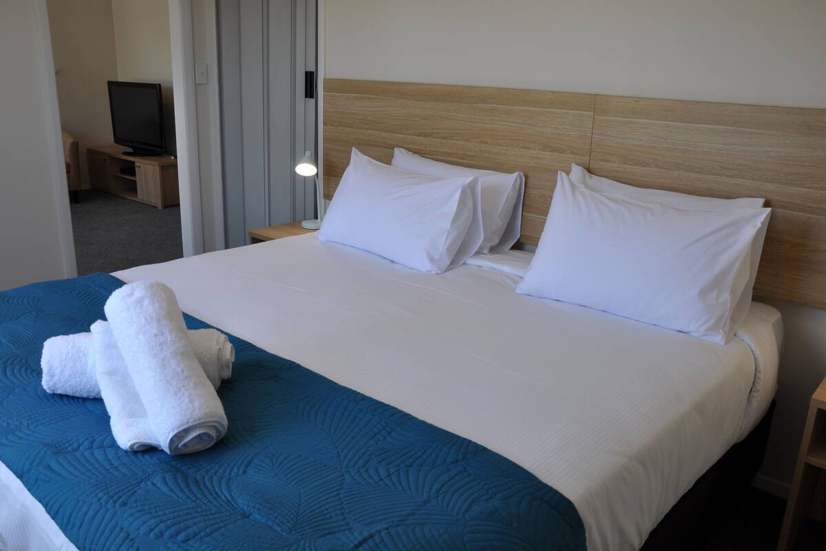 Habitación de hotel con una cama grande, ropa de cama blanca, una colcha azul decorativa y dos toallas blancas enrolladas sobre la cama.