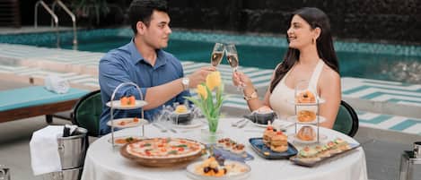 Una coppia brinda con calici di champagne a un tavolo a bordo piscina, con diversi antipasti e una piccola fontana sullo sfondo.