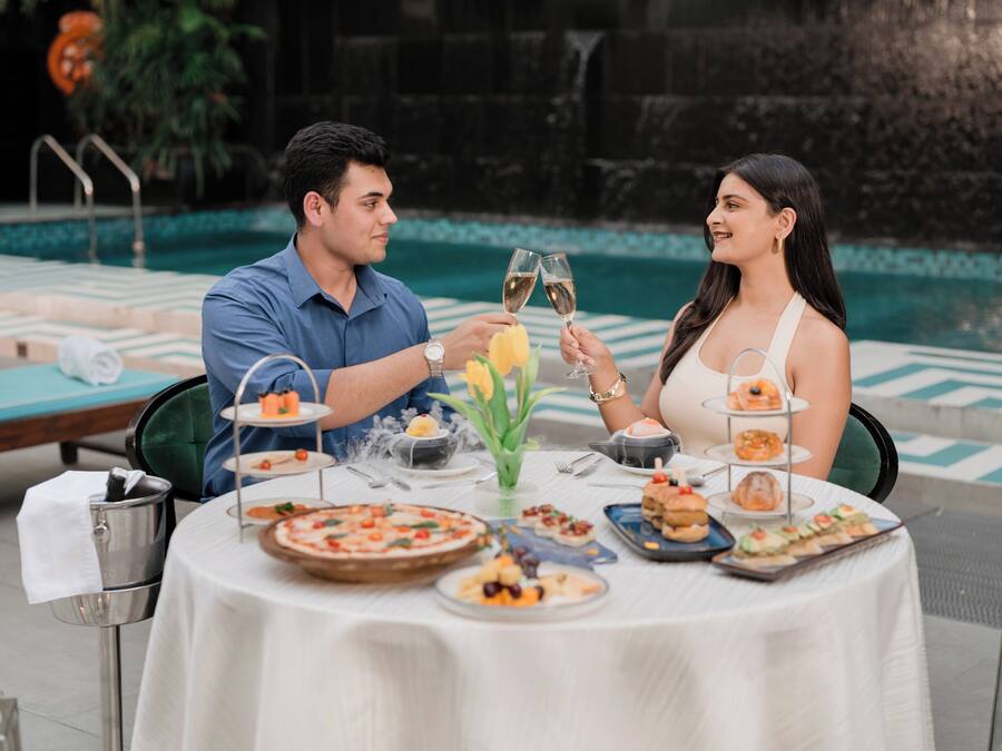 A couple toasting with champagne glasses at a poolside table with various appetizers and a small fountain in the background.