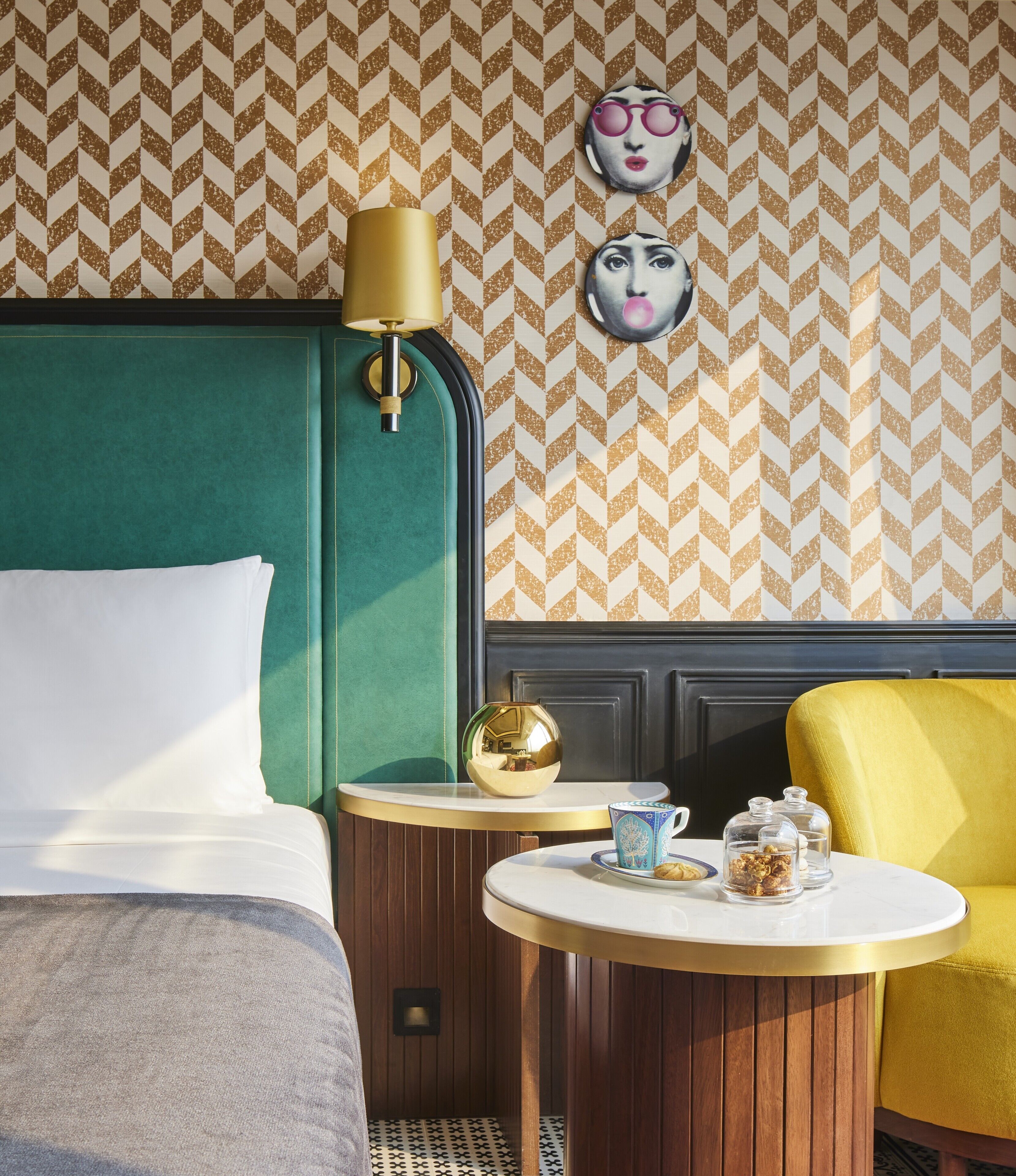 A hotel room with a green headboard, a round table with a gold lamp, and a yellow armchair.