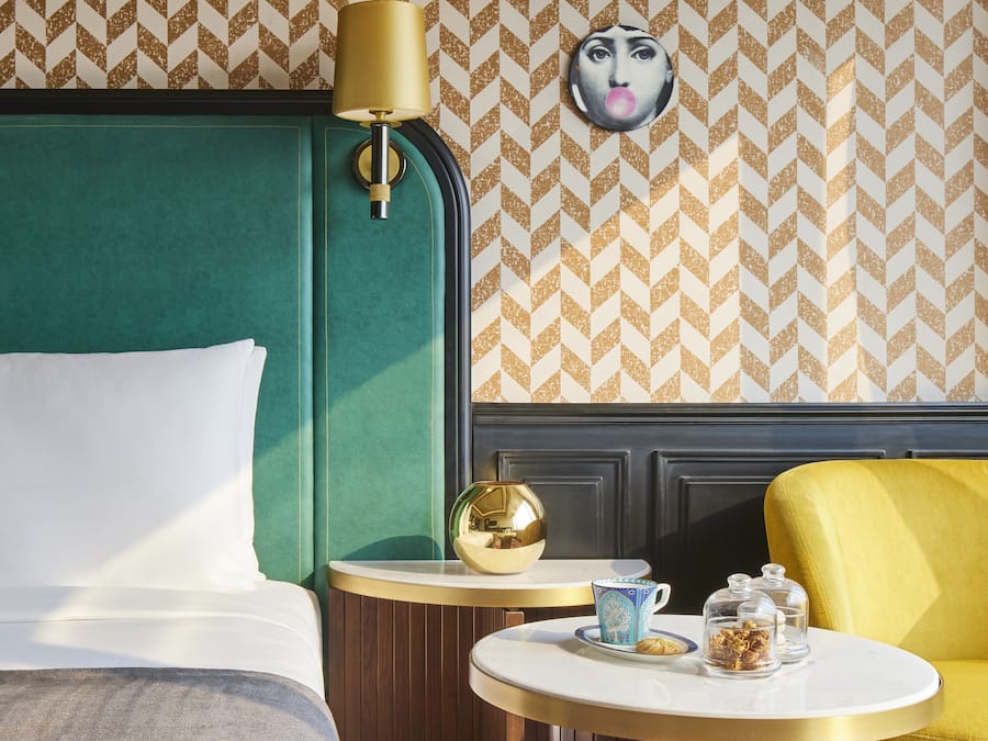 A hotel room with a green headboard, a round table with a gold lamp, and a yellow armchair.