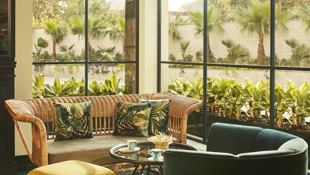 A room with a wicker sofa, a green armchair, and a round table with a tray of items. Large windows offer a view of lush greenery outside. - The Connaught, New Delhi - IHCL SeleQtions (New Delhi)