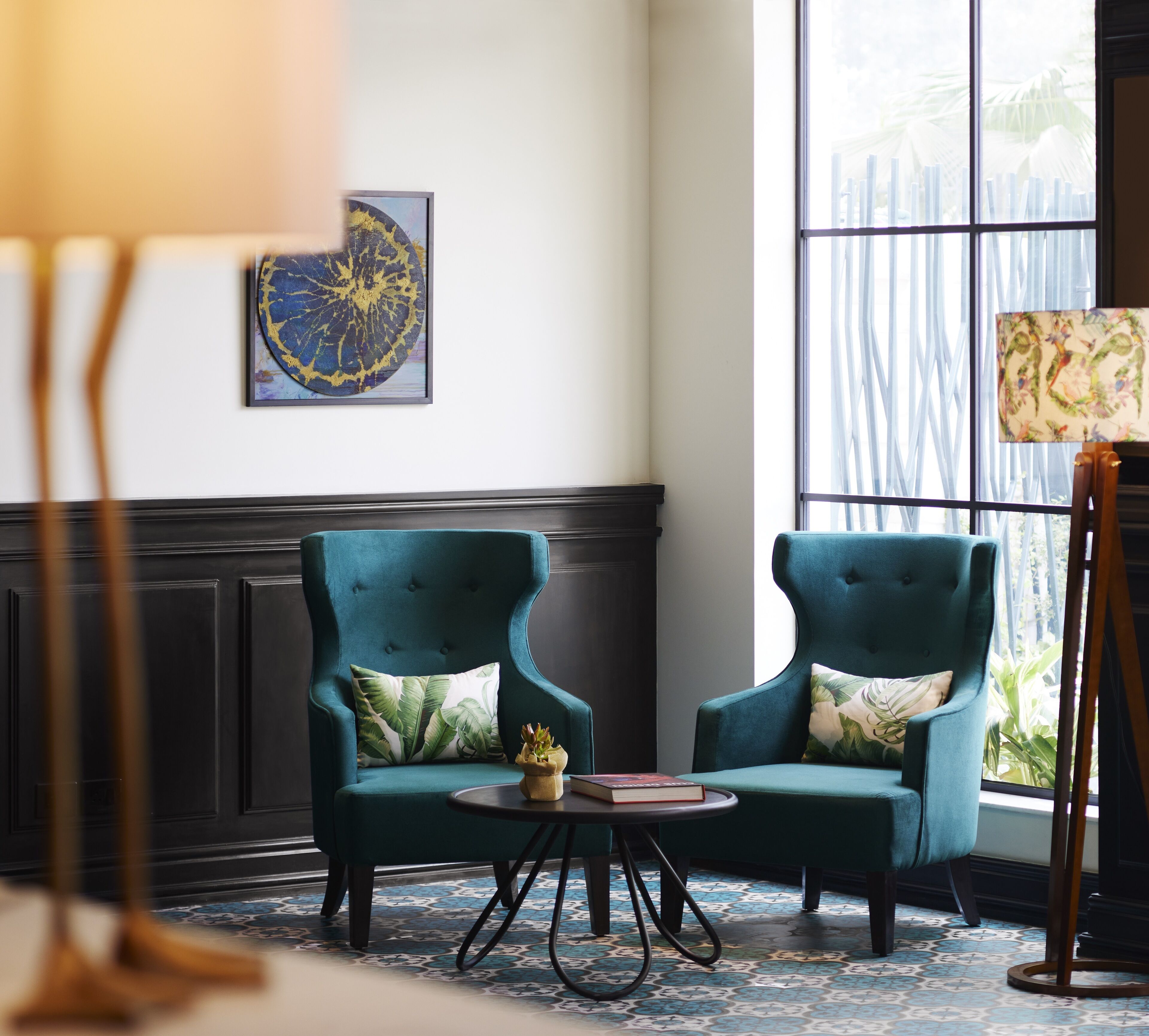 A room with two teal armchairs, a small round table with a book, and a painting on the wall.