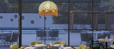 Un espace repas avec des tables rondes, des chaises recouvertes d’un tissu vert et une grande fenêtre donnant sur un espace extérieur aménagé en salon de jardin.