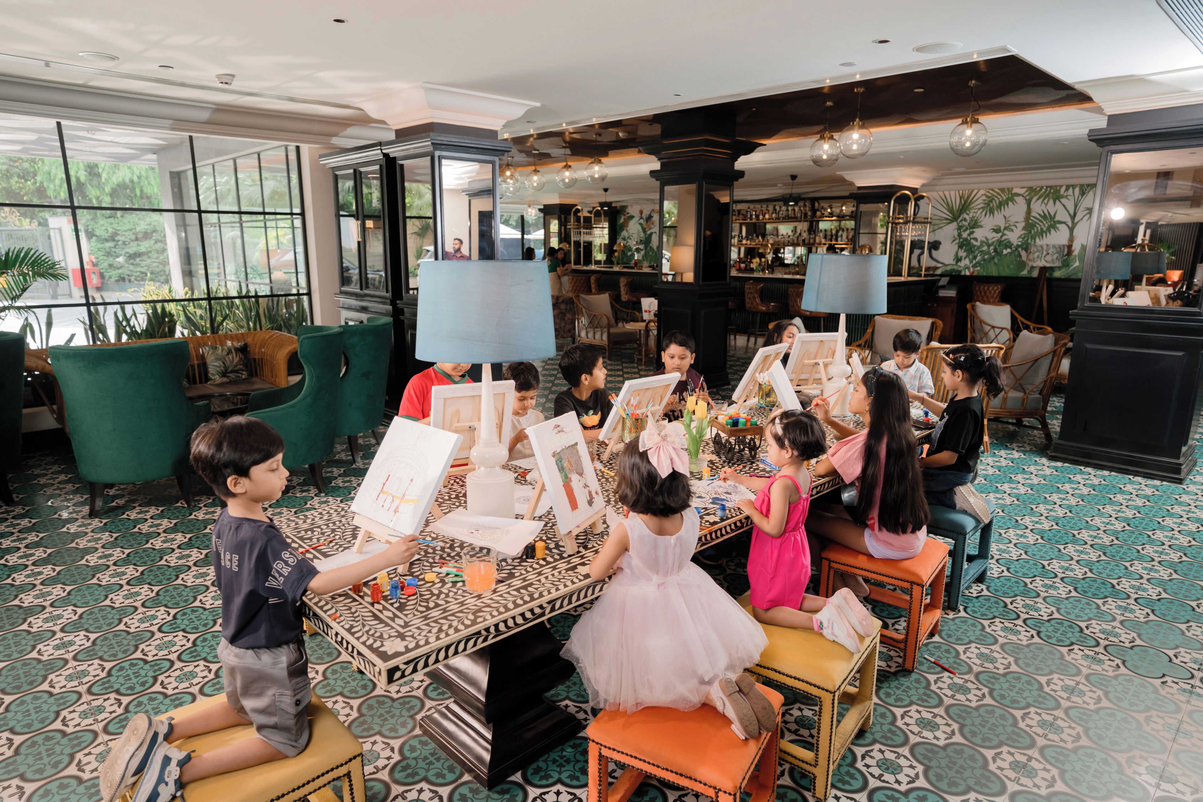 Un groupe d’enfants participait à des activités artistiques dans une pièce bien éclairée, dotée de grandes fenêtres, d’un bar et d’un sol à motifs.