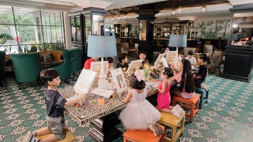 Un groupe d’enfants participait à des activités artistiques dans une pièce bien éclairée, dotée de grandes fenêtres, d’un bar et d’un sol à motifs.