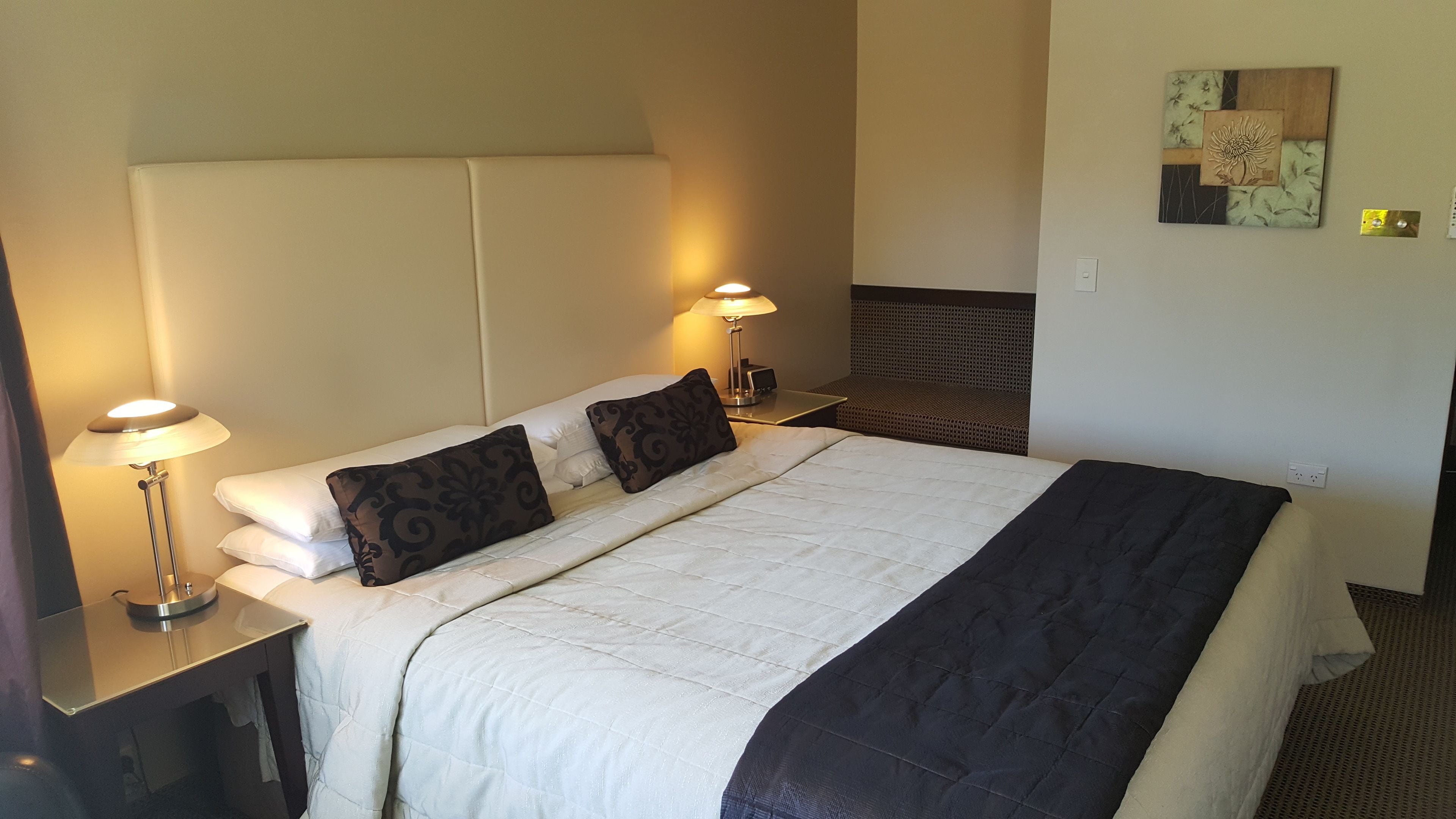 A hotel room with a large bed, two bedside tables, a nightstand, a wall-mounted lamp, and a decorative piece on the wall.