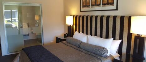 A hotel room with a bed, bedside lamps, a framed picture of barrels, and a bathroom visible through a glass door.