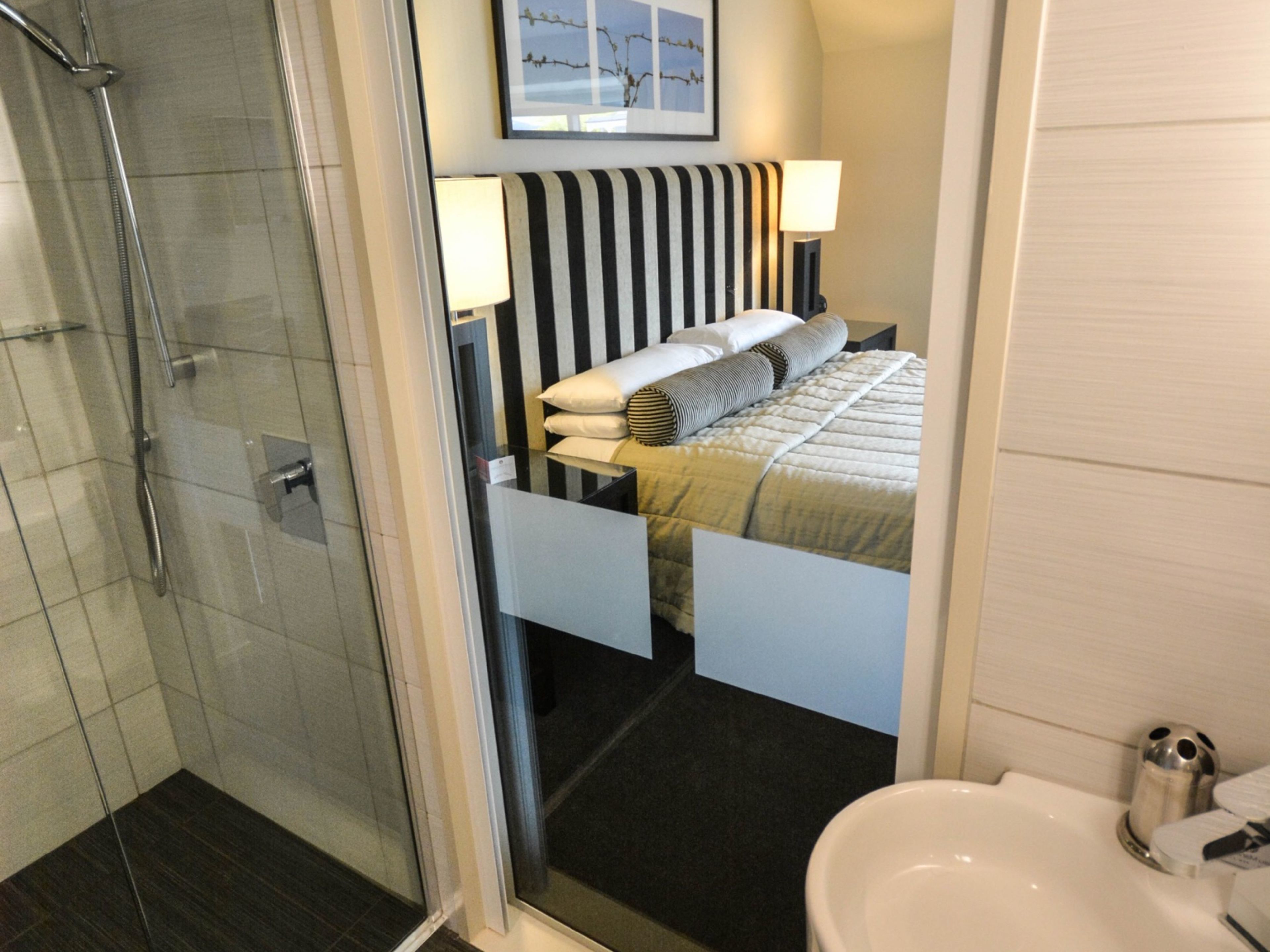 Habitació d'hotel amb una dutxa de vidre, un llit amb roba de llit estriada, una nit de nit amb una làmpada i un quadre encimbellat a la paret.