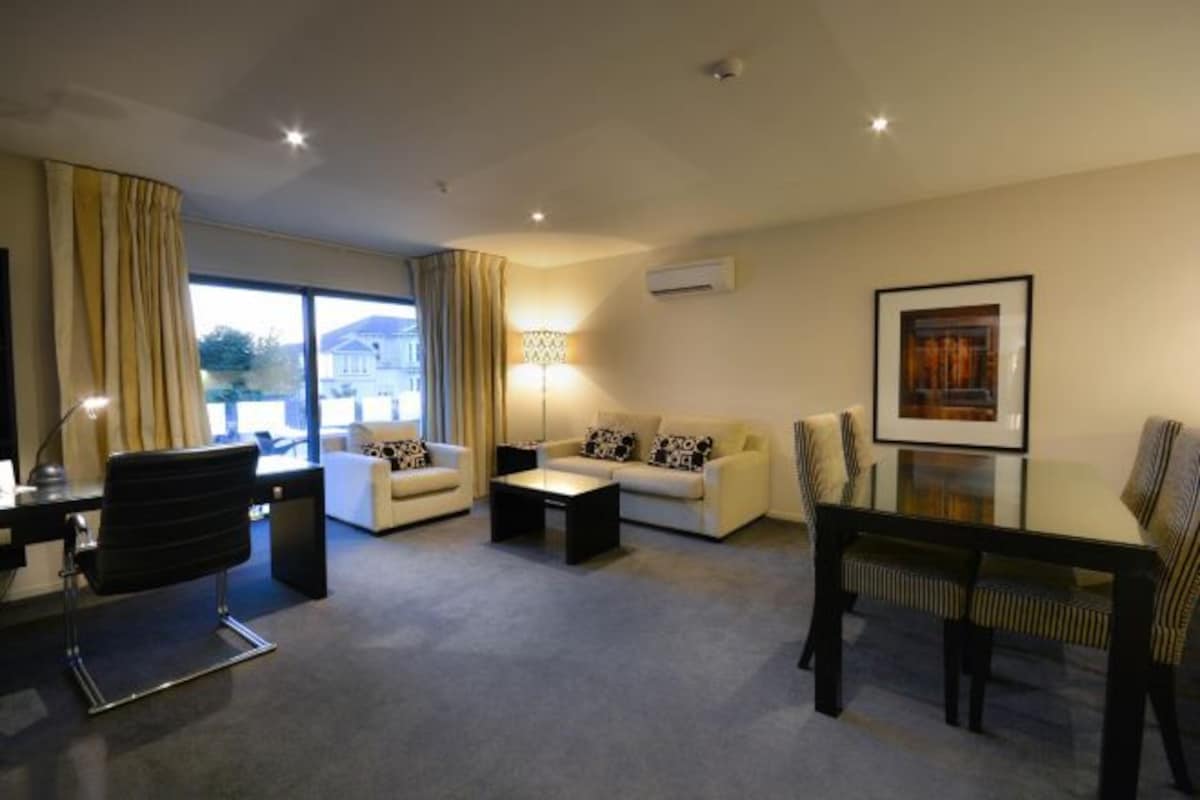 A modern living room with a sofa, coffee table, and a dining table with chairs. There is a large window with curtains, an air conditioning unit on the wall, and a framed picture on the wall.