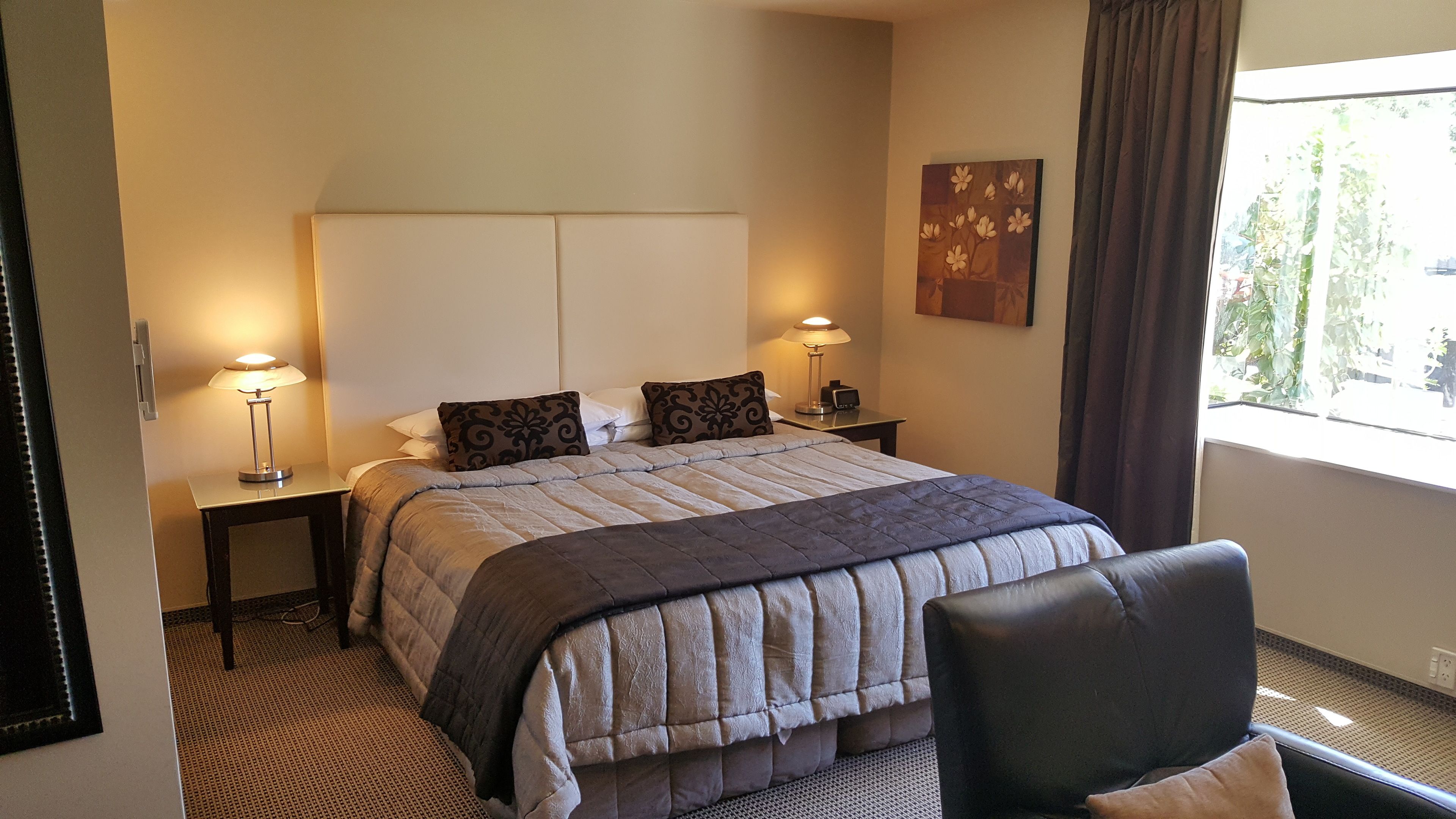 Une chambre d’hôtel comprenant un lit, deux lampes de chevet, une chaise, une fenêtre avec des rideaux et une œuvre d’art accrochée au mur.