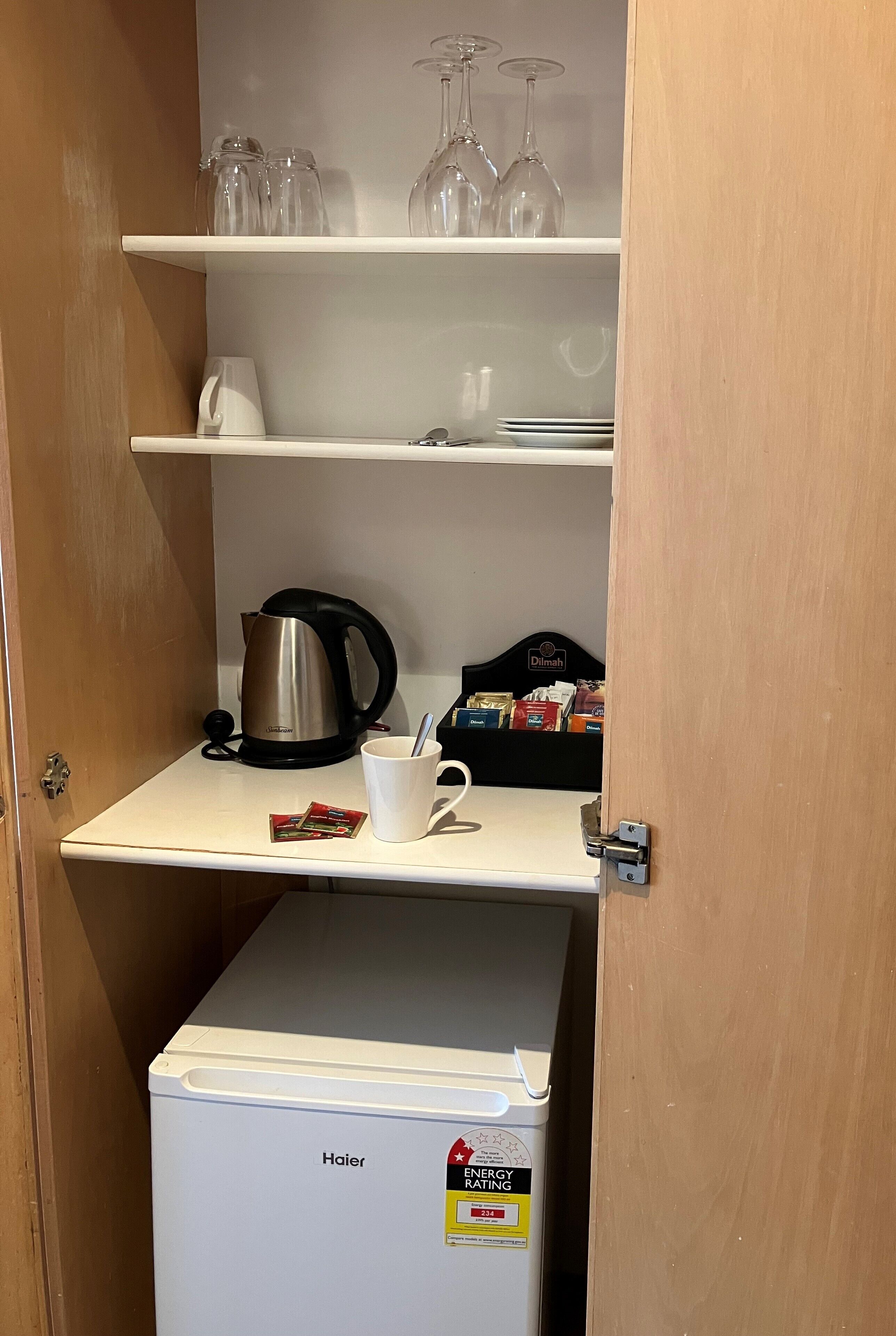 A compact kitchenette with a Haier refrigerator, a coffee maker, a kettle, and a shelf with glassware and a box of tea.