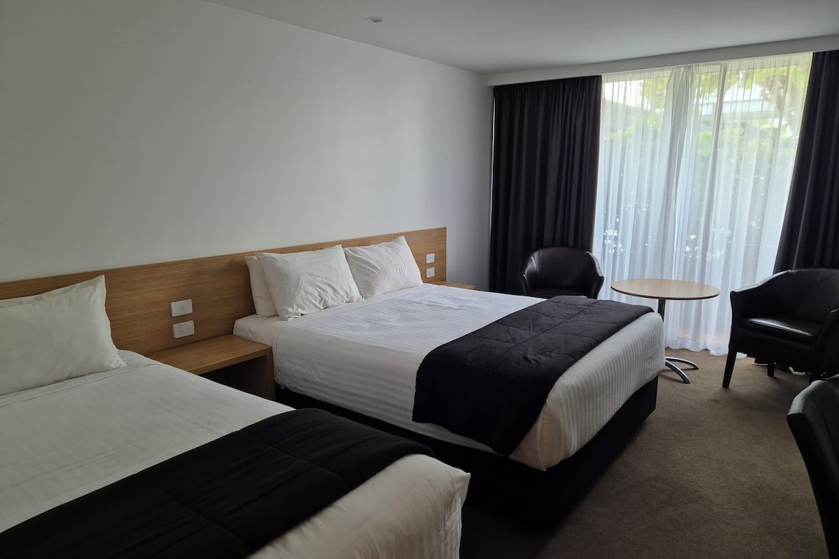 A hotel room with two beds, a small table, a chair, and a window with curtains.